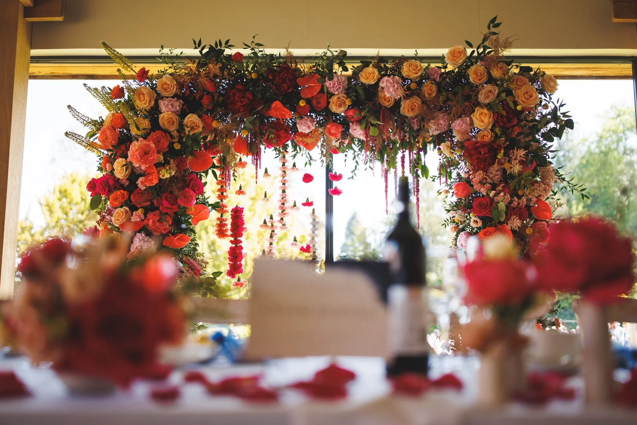 Colorful floral arch with pink, red, and peach flowers, set against a large window with outdoor greenery in the background.