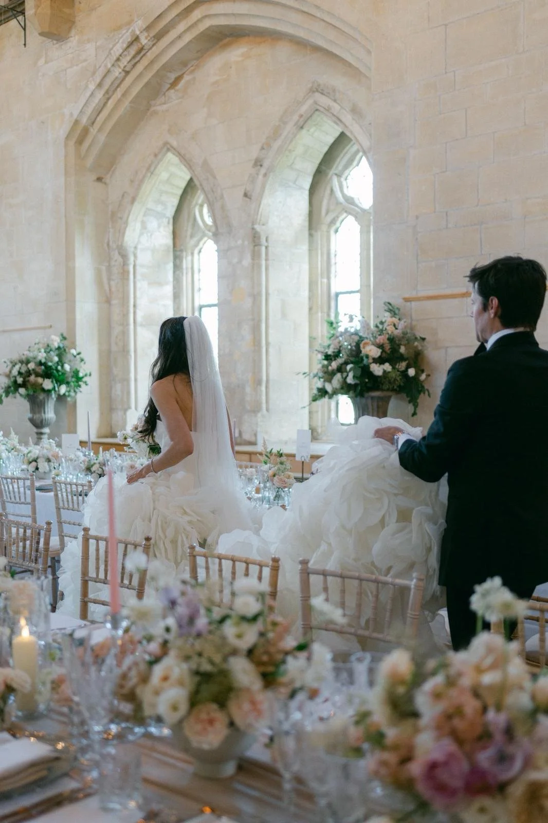 A bride and groom at their wedding reception in a church with stone walls and high arched windows, decorated with floral arrangements and a elegantly set table in the foreground.