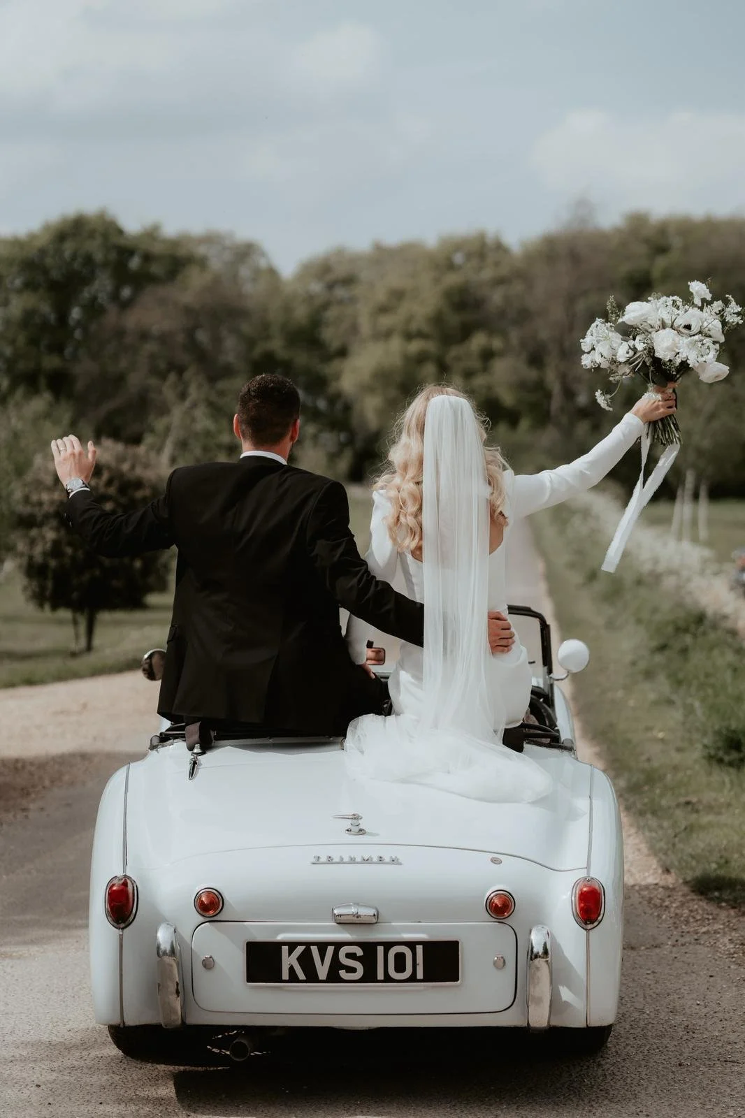 A bride and groom riding in a vintage white car on a country road during their wedding celebration, with the bride holding a bouquet of white flowers and all four people dressed formally.