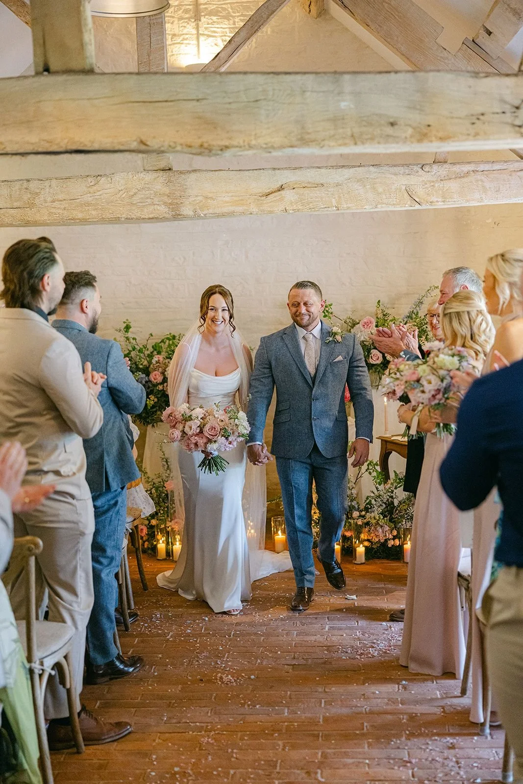 Bride and groom holding hands walking down aisle during wedding ceremony, surrounded by guests clapping and smiling, decorated with candles and flowers.