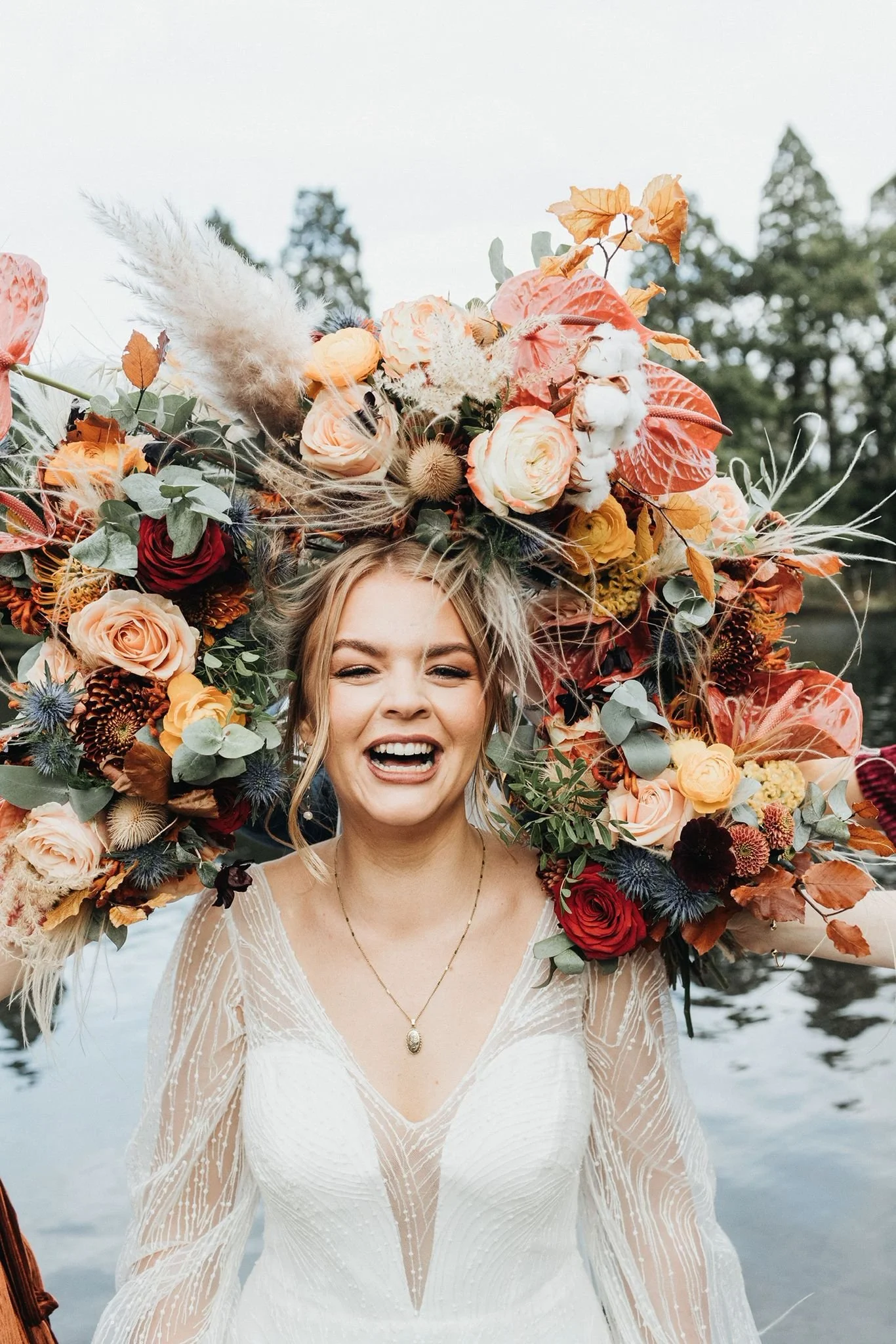 A woman in a white dress with a large floral headpiece made of various colorful flowers and autumn leaves, standing outdoors near water with trees in the background.
