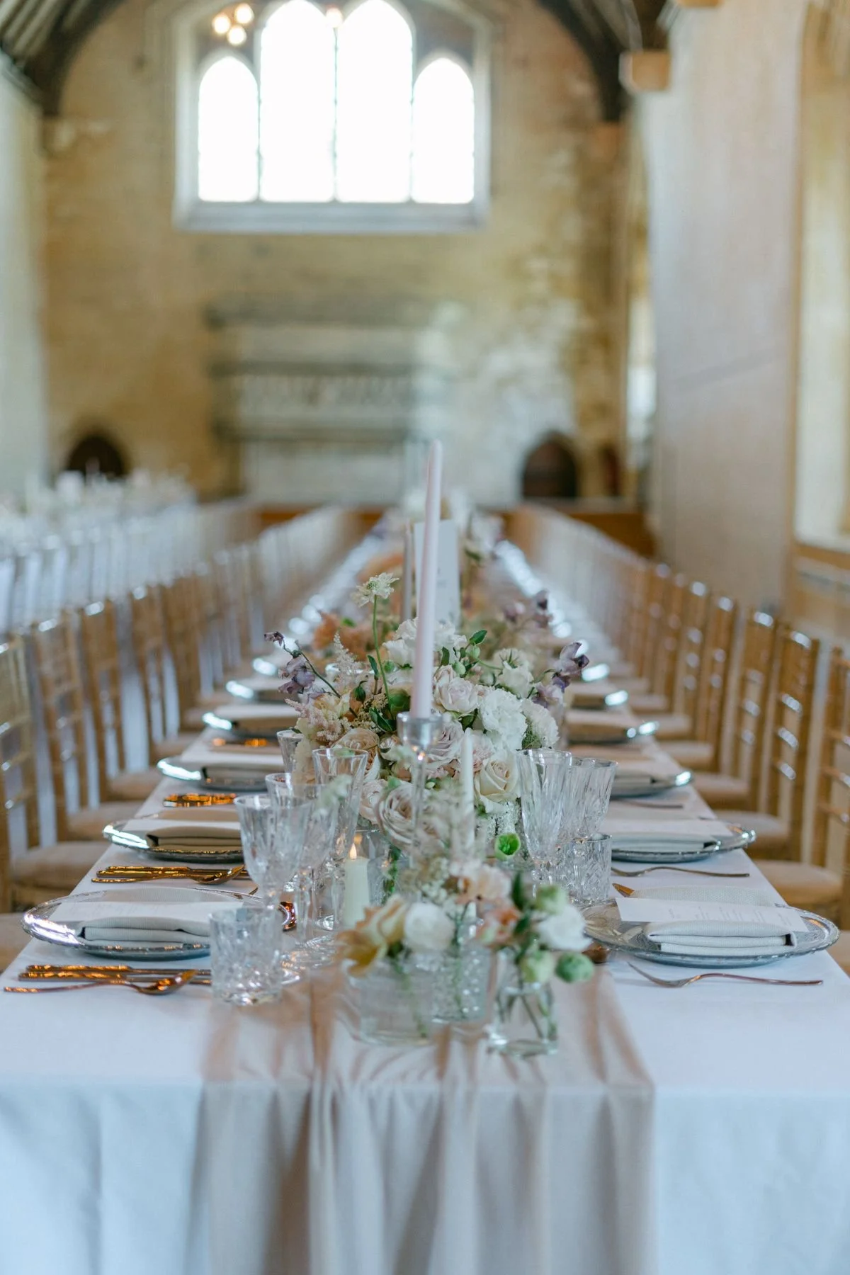 Elegant dining table set for a wedding with white floral arrangements, candles, and glassware in a rustic stone hall.