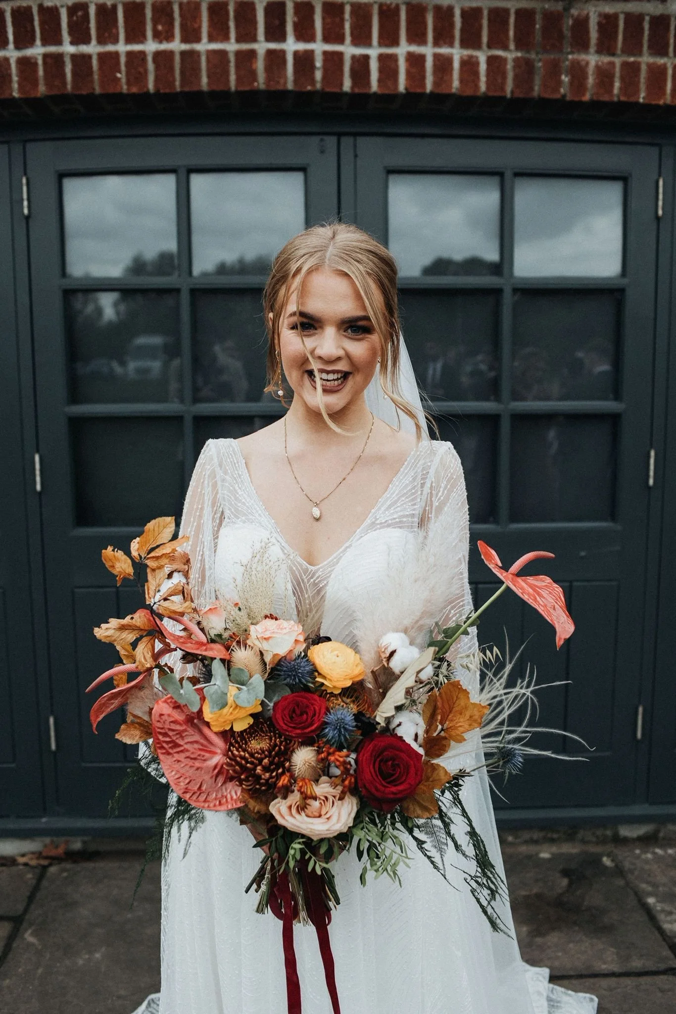 A smiling bride in a white wedding dress holding a colorful bouquet of flowers in front of a dark gray door and brick wall.