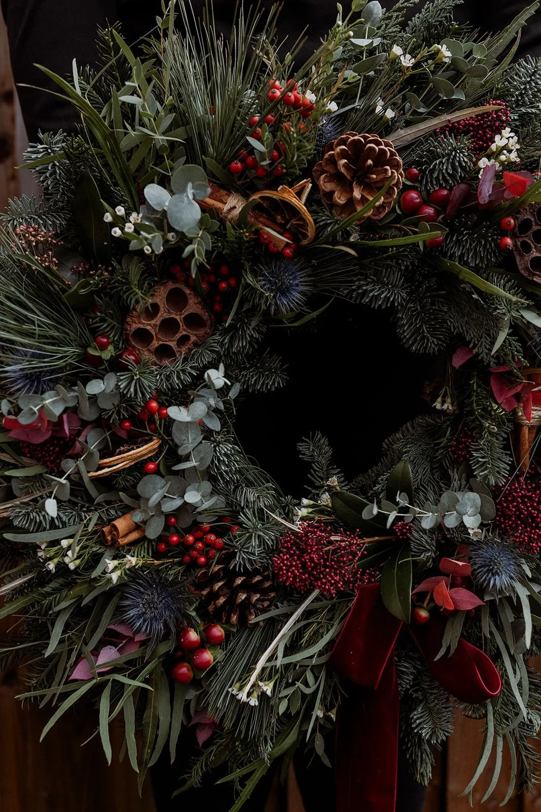 Close-up of a Christmas wreath made of pine, eucalyptus, berries, pinecones, cinnamon sticks, dried flowers, and red ribbon.