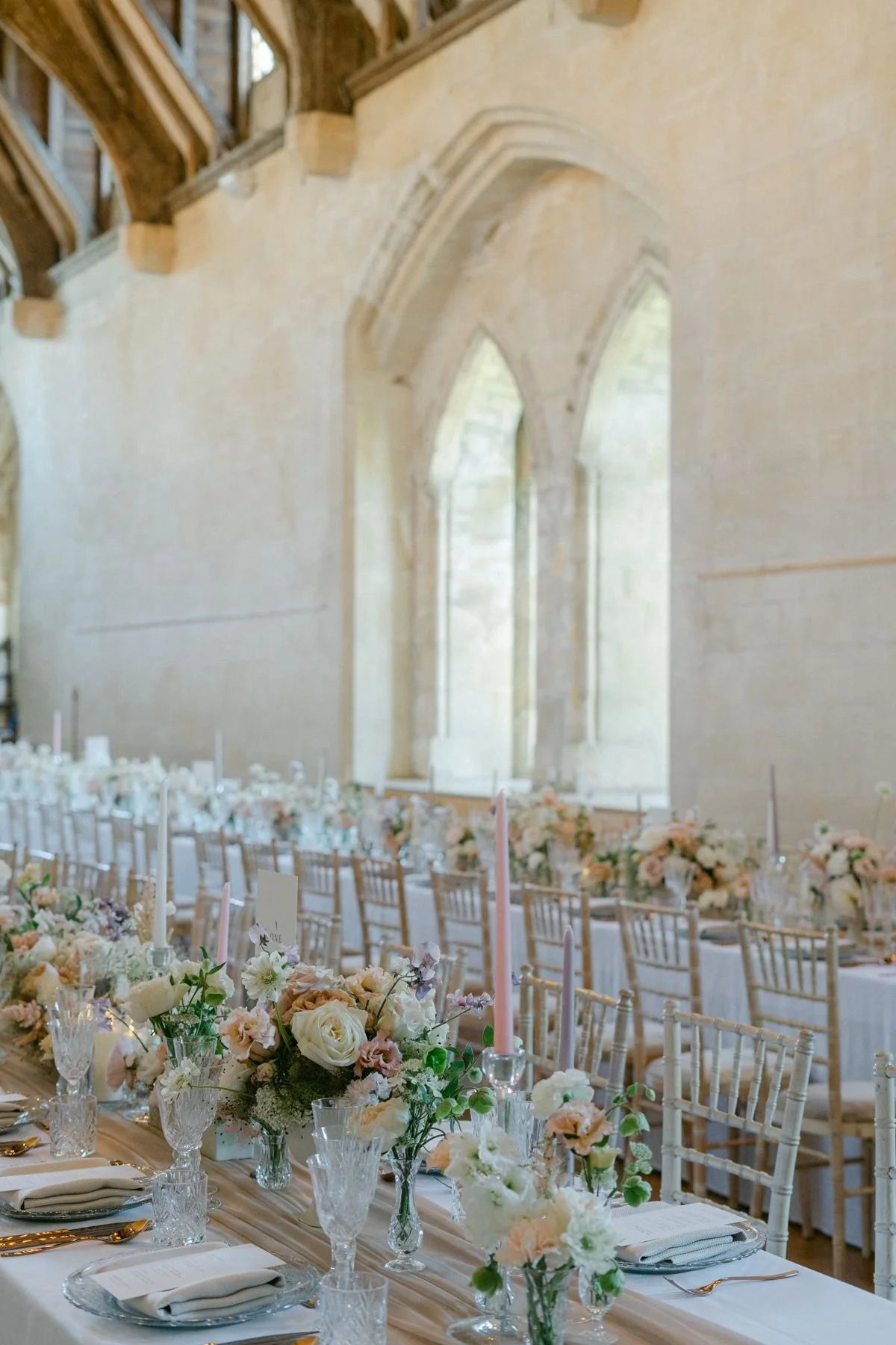 Beautifully decorated banquet table with floral centerpieces, glassware, and napkins inside a stone hall with arched windows.