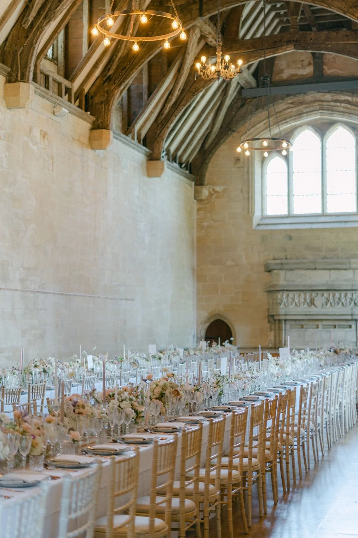 Elegant banquet hall with a long table decorated with flower arrangements, set with plates, silverware, and glasses. The hall features stone walls, large windows, and a high, wooden, vaulted ceiling with chandeliers and circular light fixtures.