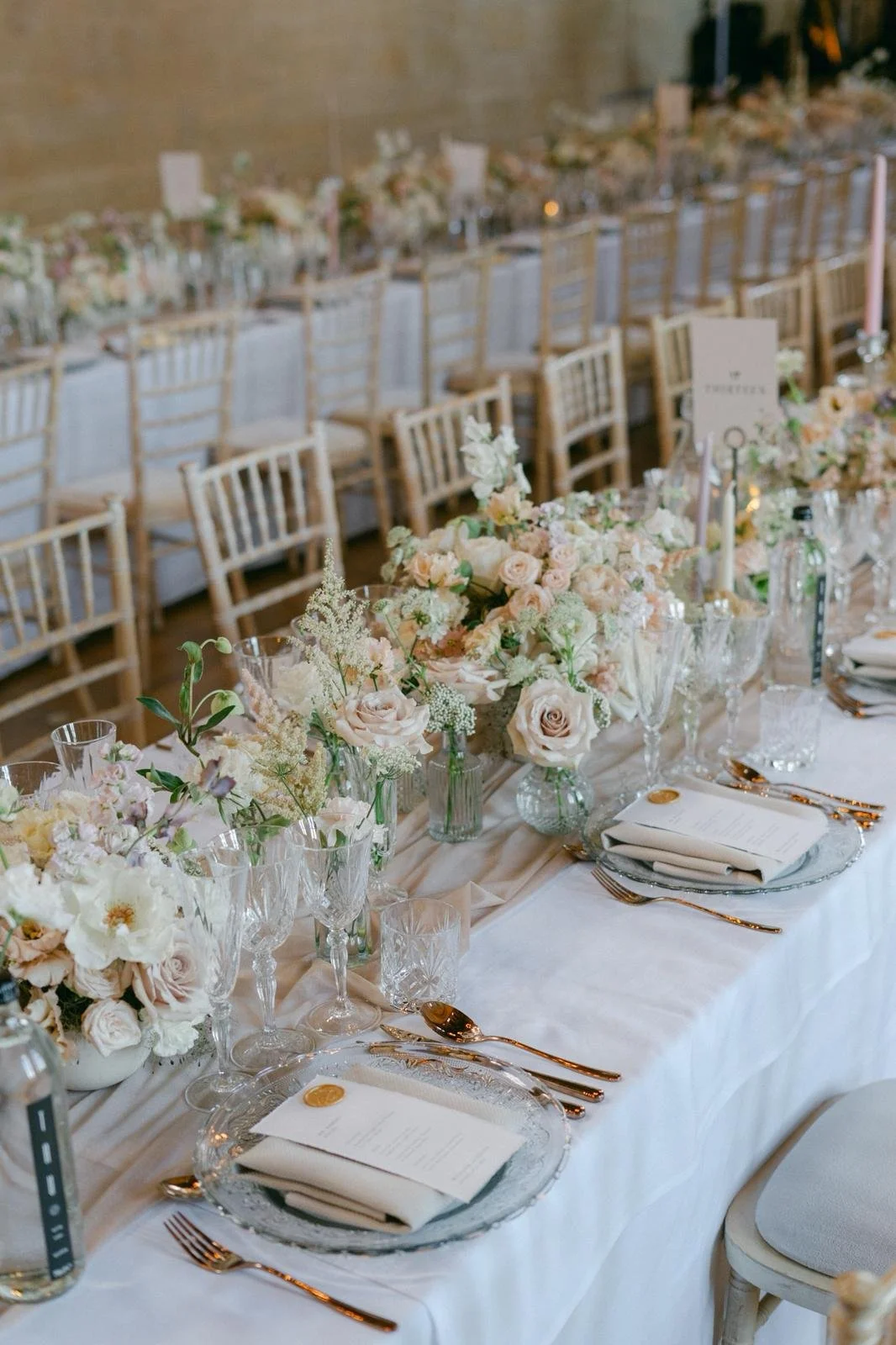 Elegant wedding reception table decorated with pink and white flowers in glass vases, set with glassware, gold flatware, and white napkins, with beige chairs in the background.