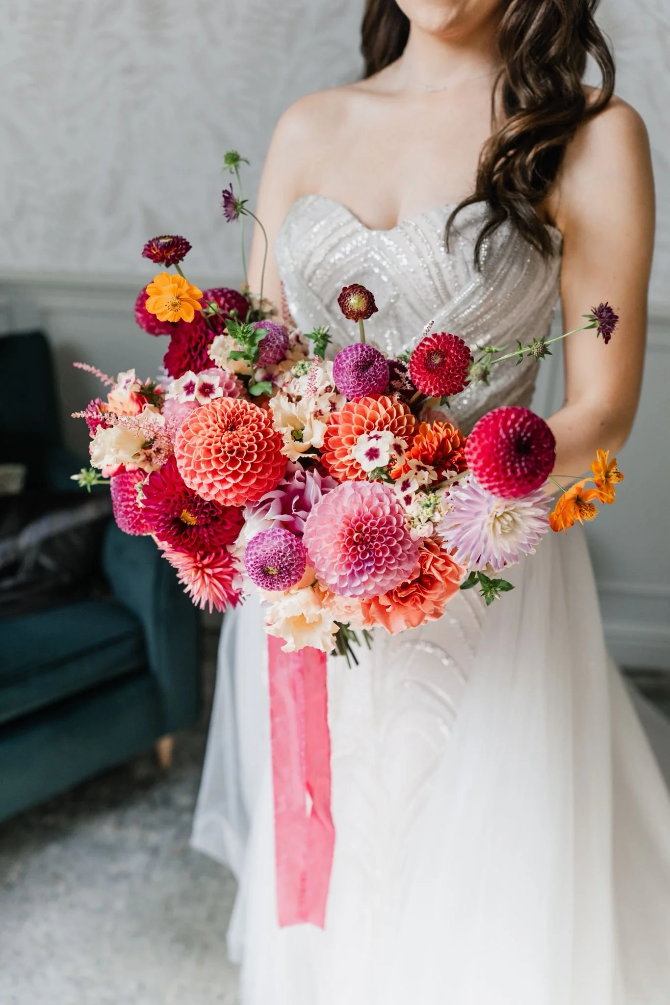 A woman in a wedding dress holding a large bouquet of colorful flowers, including dahlias, scaevola, and zinnias, with a pink ribbon hanging from the bouquet.