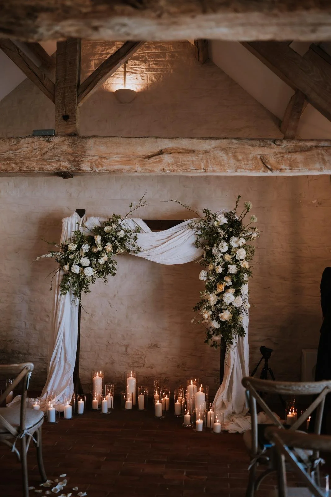 Wedding altar decorated with white flowers and draped white fabric, surrounded by lit candles on the floor, in a rustic indoor venue with exposed wooden beams and brick walls.