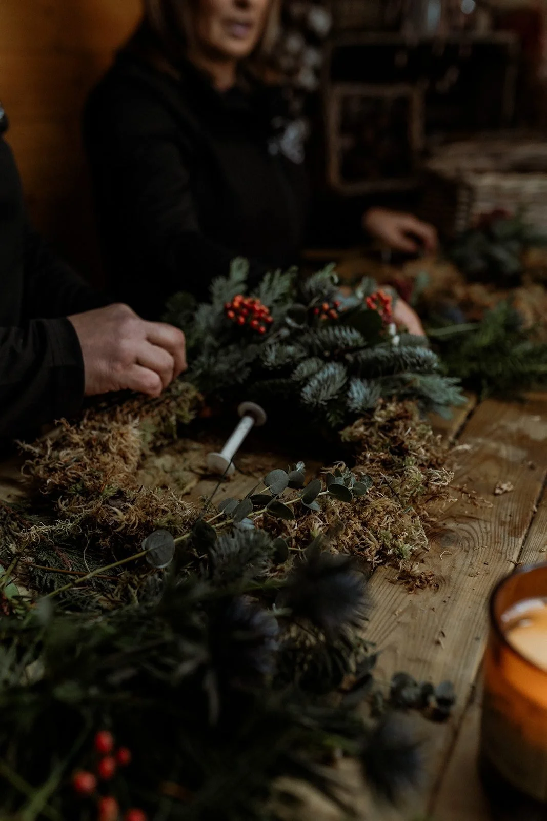 People arranging holiday greenery and decorations on a wooden table, with supplies and candles nearby.