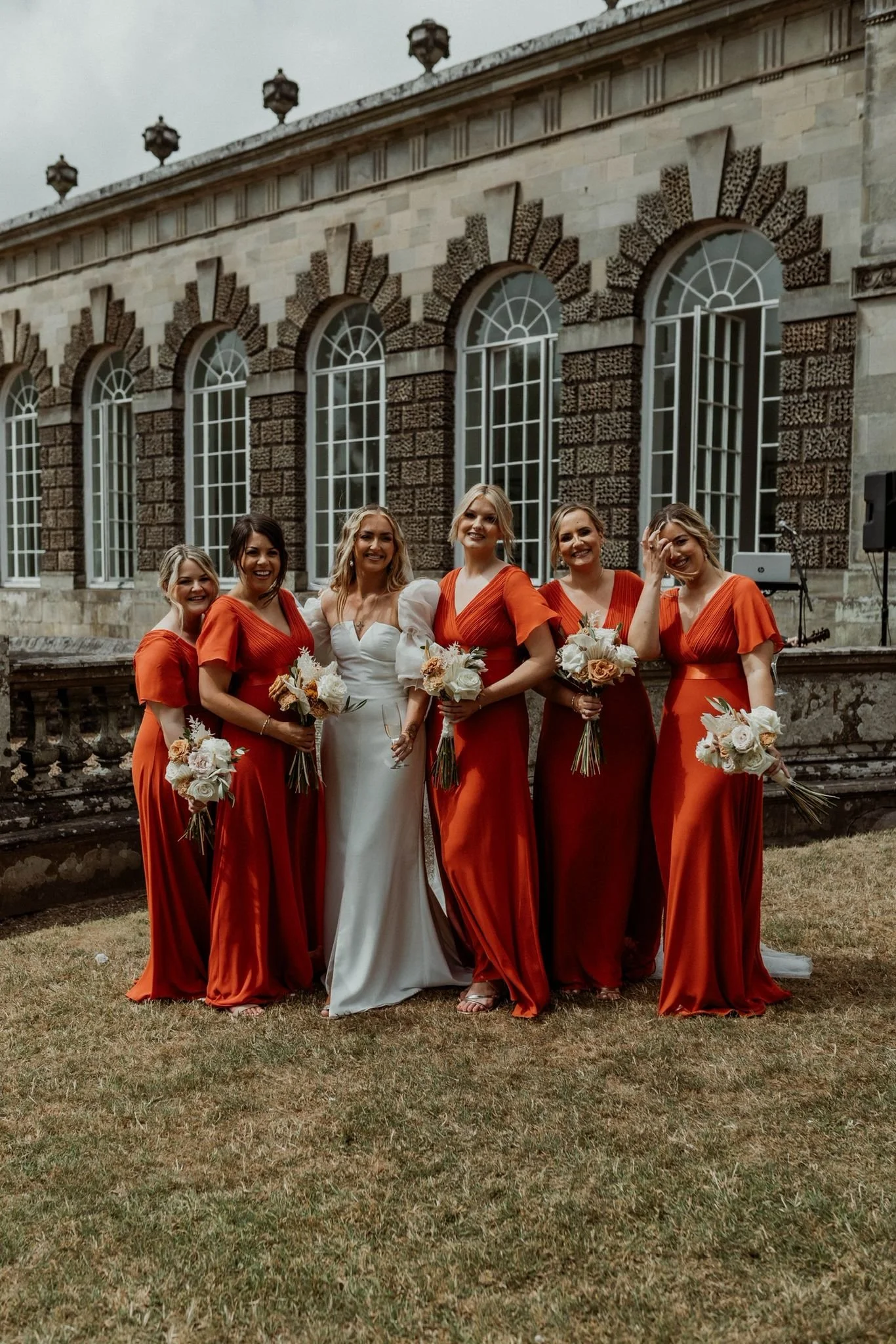 A group of six women, five bridesmaids in orange dresses and one bride in a white gown, standing outdoors in front of a historic building with large arched windows, holding bouquets of flowers, smiling at the camera.