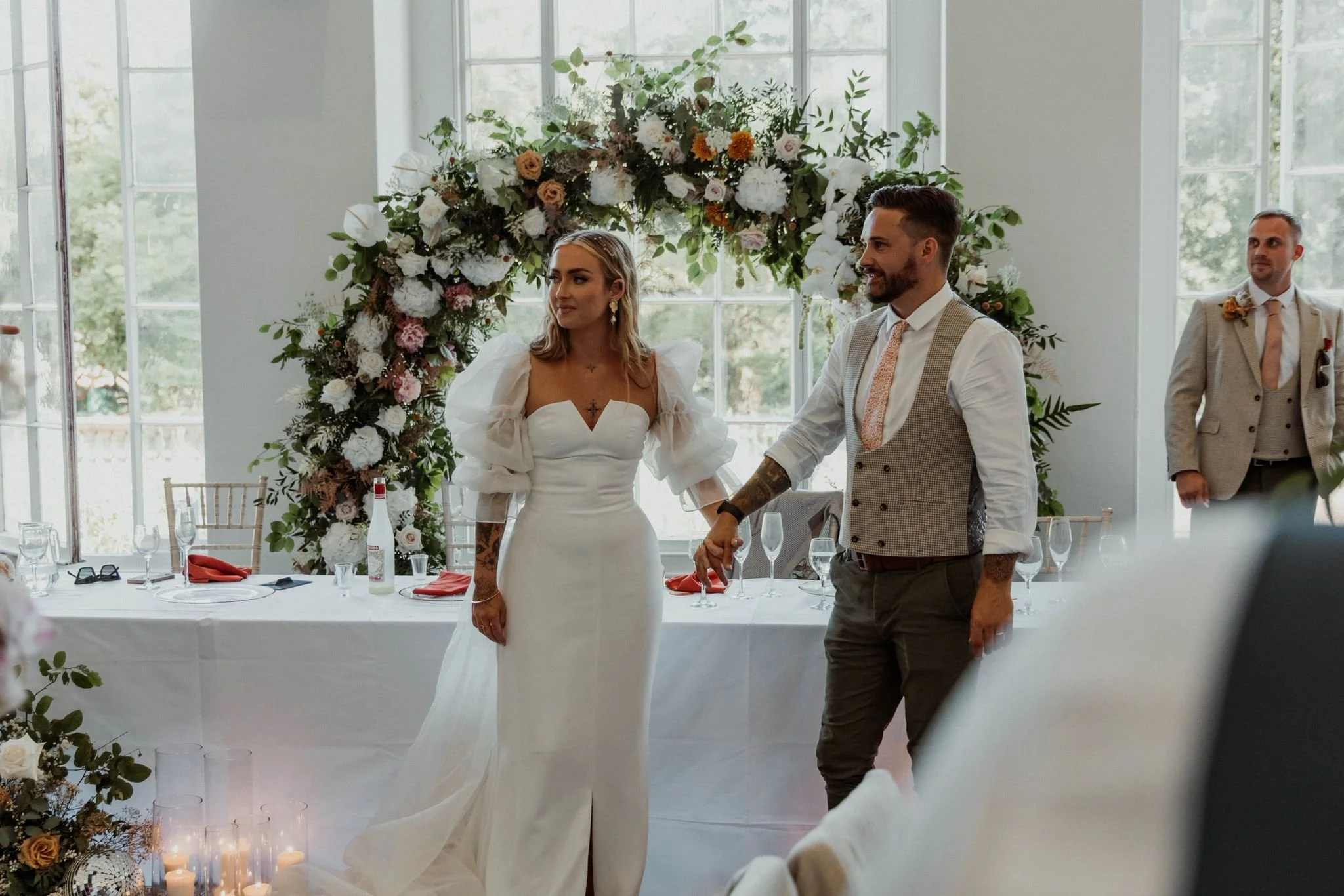 A bride and groom holding hands during their wedding ceremony, with an officiant in the background, in front of a floral arch in a bright, indoor setting.