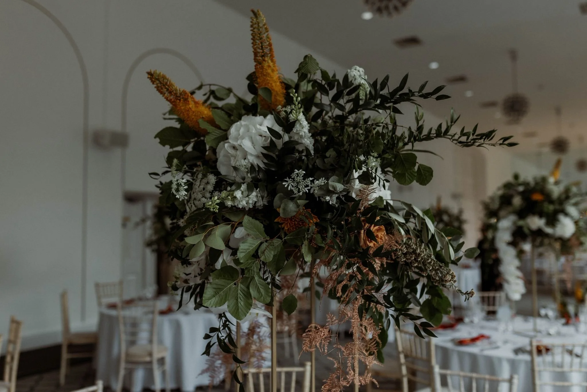 Elegant floral arrangement with white and orange flowers, green leaves, set in a decorated event space with round tables and chairs.