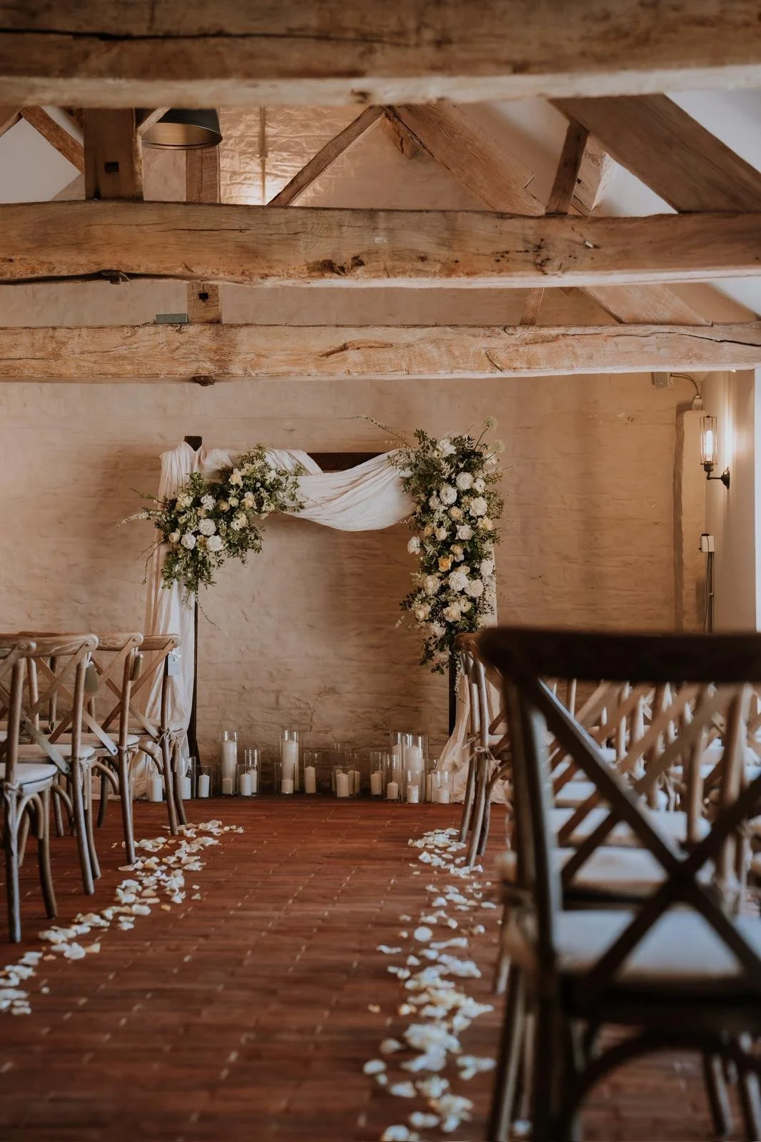Indoor wedding ceremony setup with wooden chairs lining a brick aisle, white flower petal decorations, and an arch decorated with white flowers and drapery.