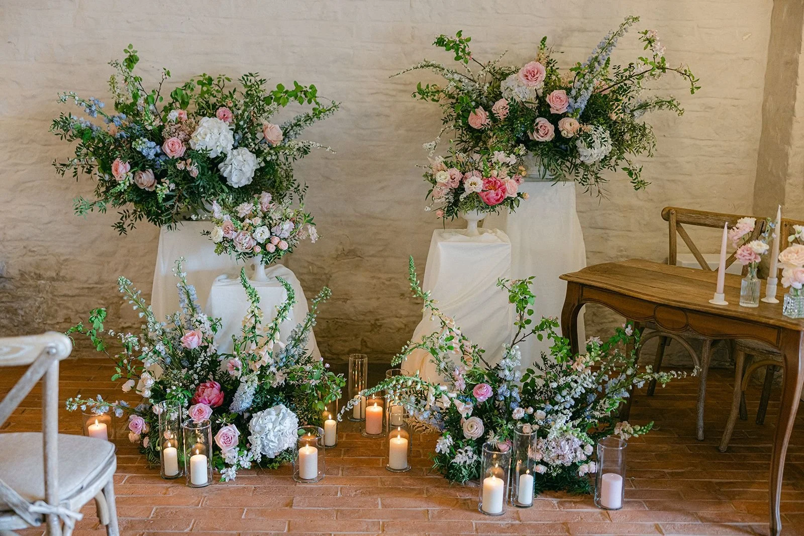 Elegant floral arrangements with pink, white, and lavender flowers, accented by candles on the floor, set against a textured wall, possibly for a wedding or special event.