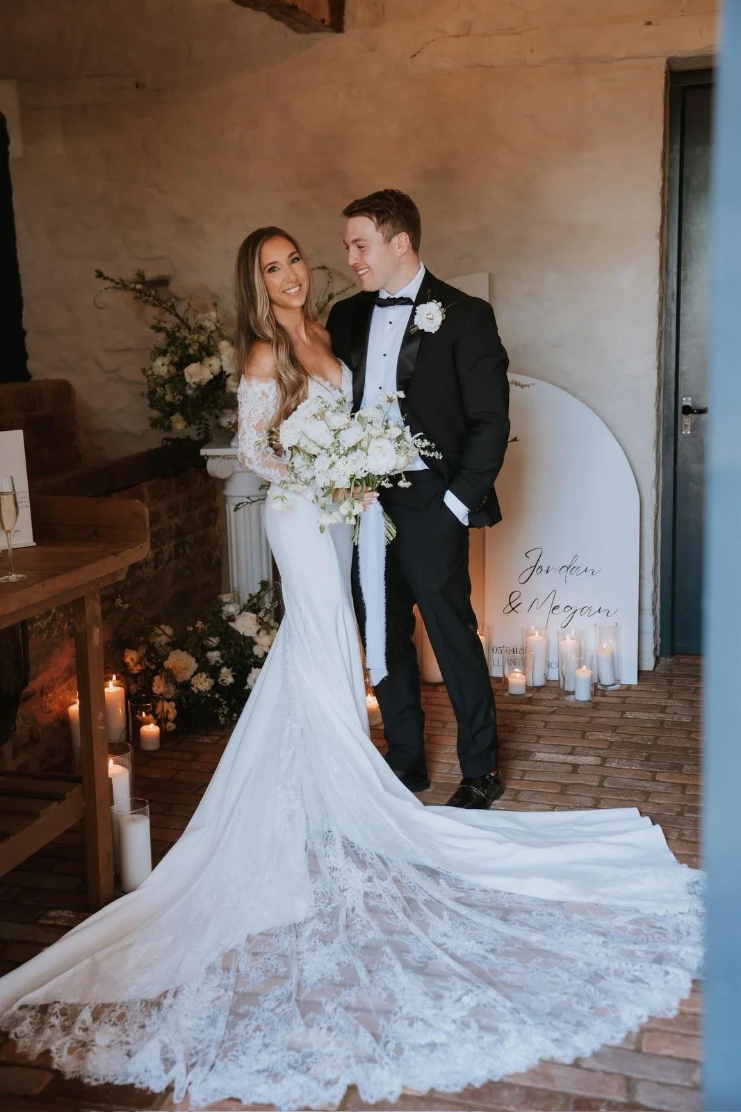 A bride and groom stand together indoors on their wedding day, smiling and holding a bouquet of white flowers. The bride wears a long white lace wedding gown with a train, and the groom wears a black tuxedo with a bow tie. Candles and floral decor ar