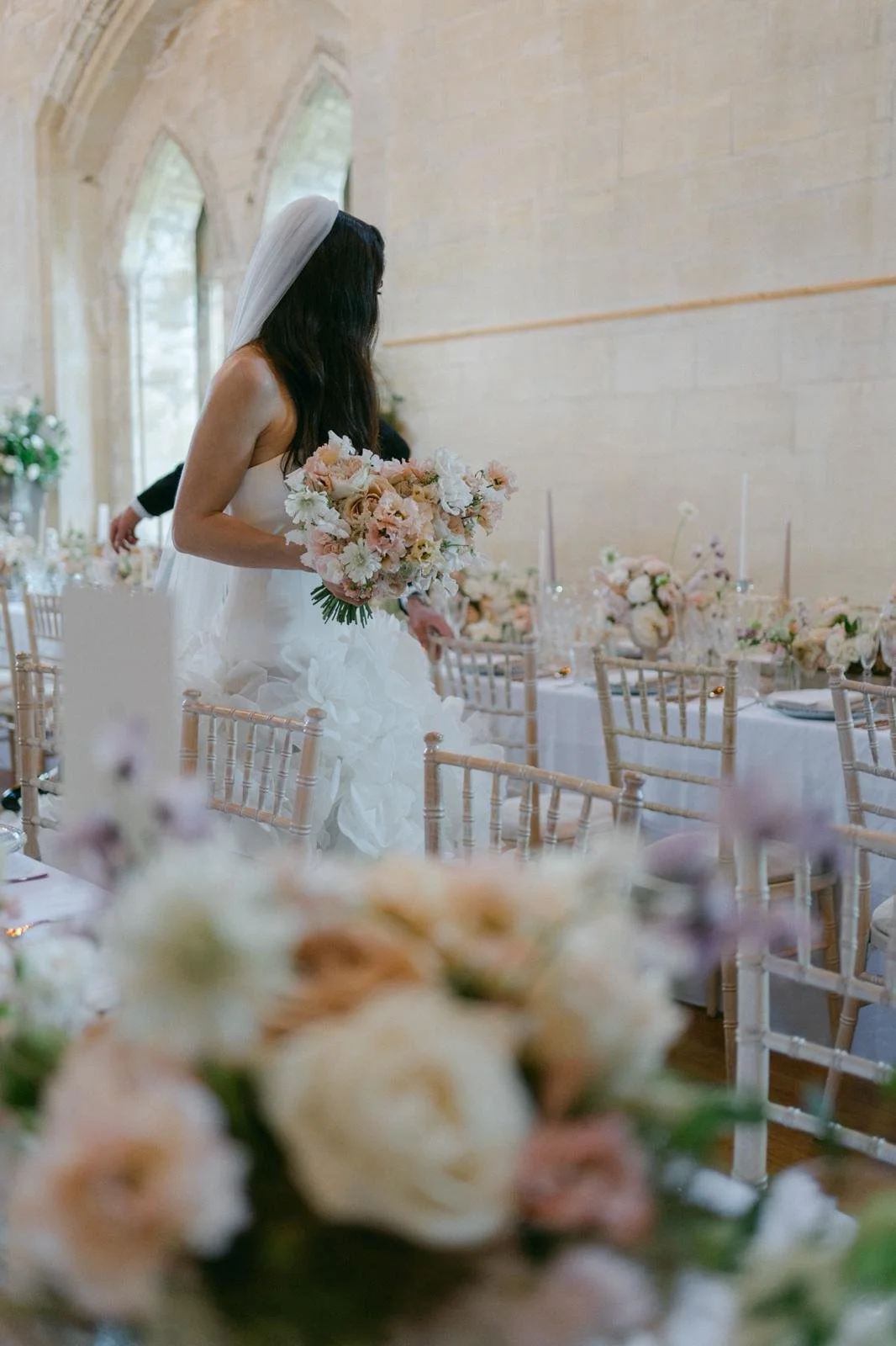 Bride in wedding dress holding a bouquet of white and blush flowers, standing among decorated tables with floral arrangements in a church or hall with arched windows.
