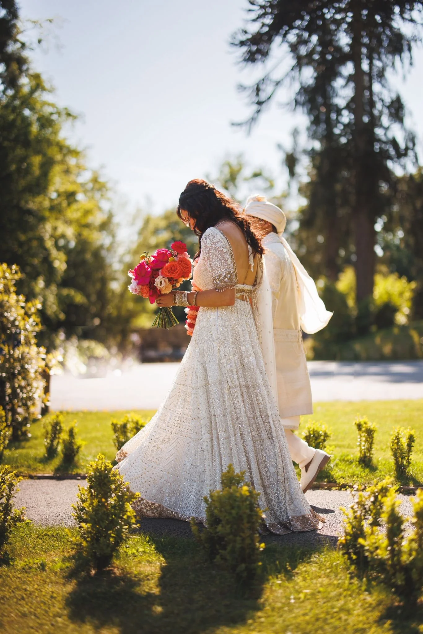 A bride in a white wedding dress holding a bouquet of pink, red, orange, and cream flowers, with an outdoor garden in the background.