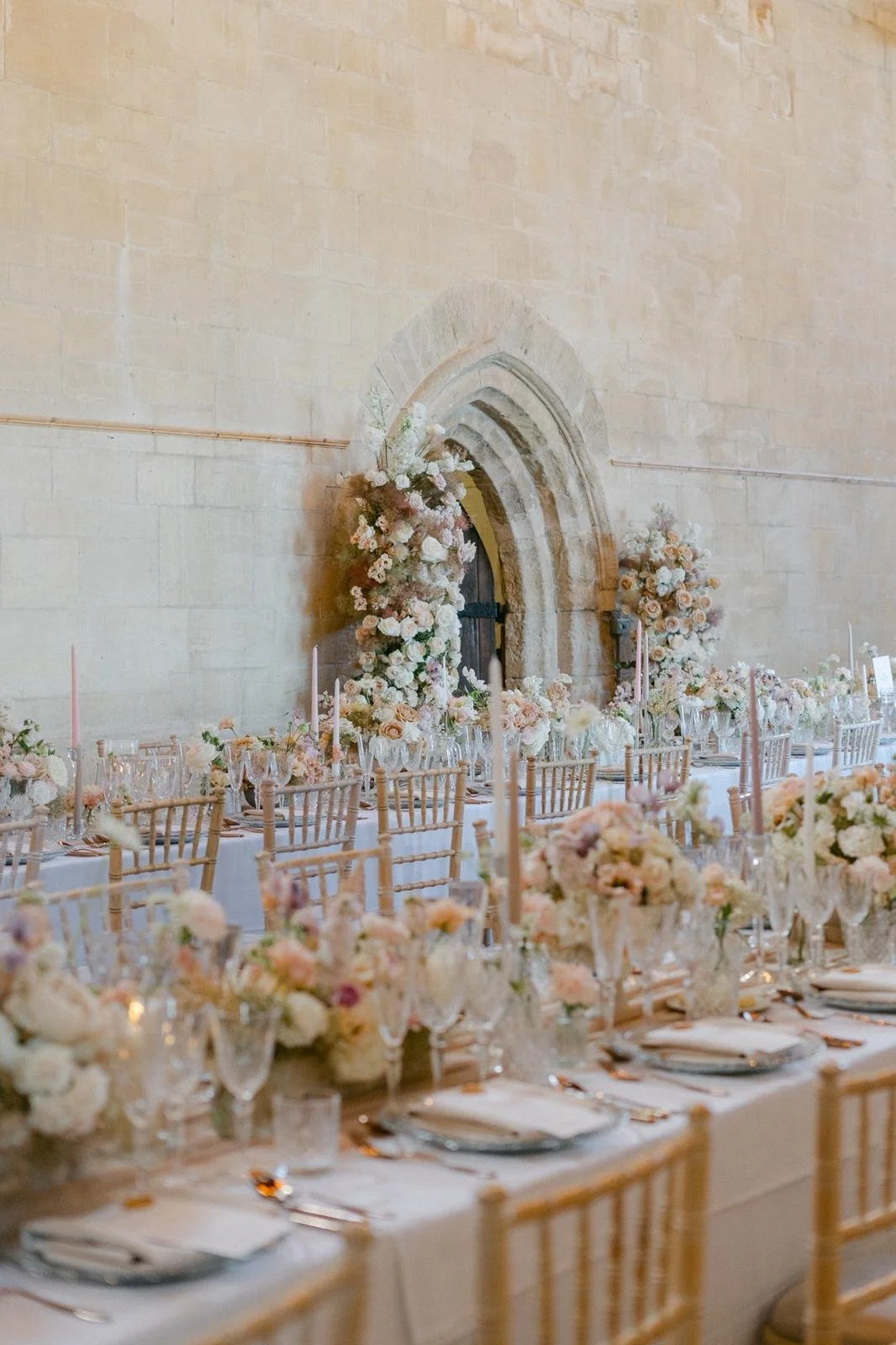 Wedding reception table with floral arrangements, candles, glassware, and place settings, set against a stone wall with an arched window and floral decorations.