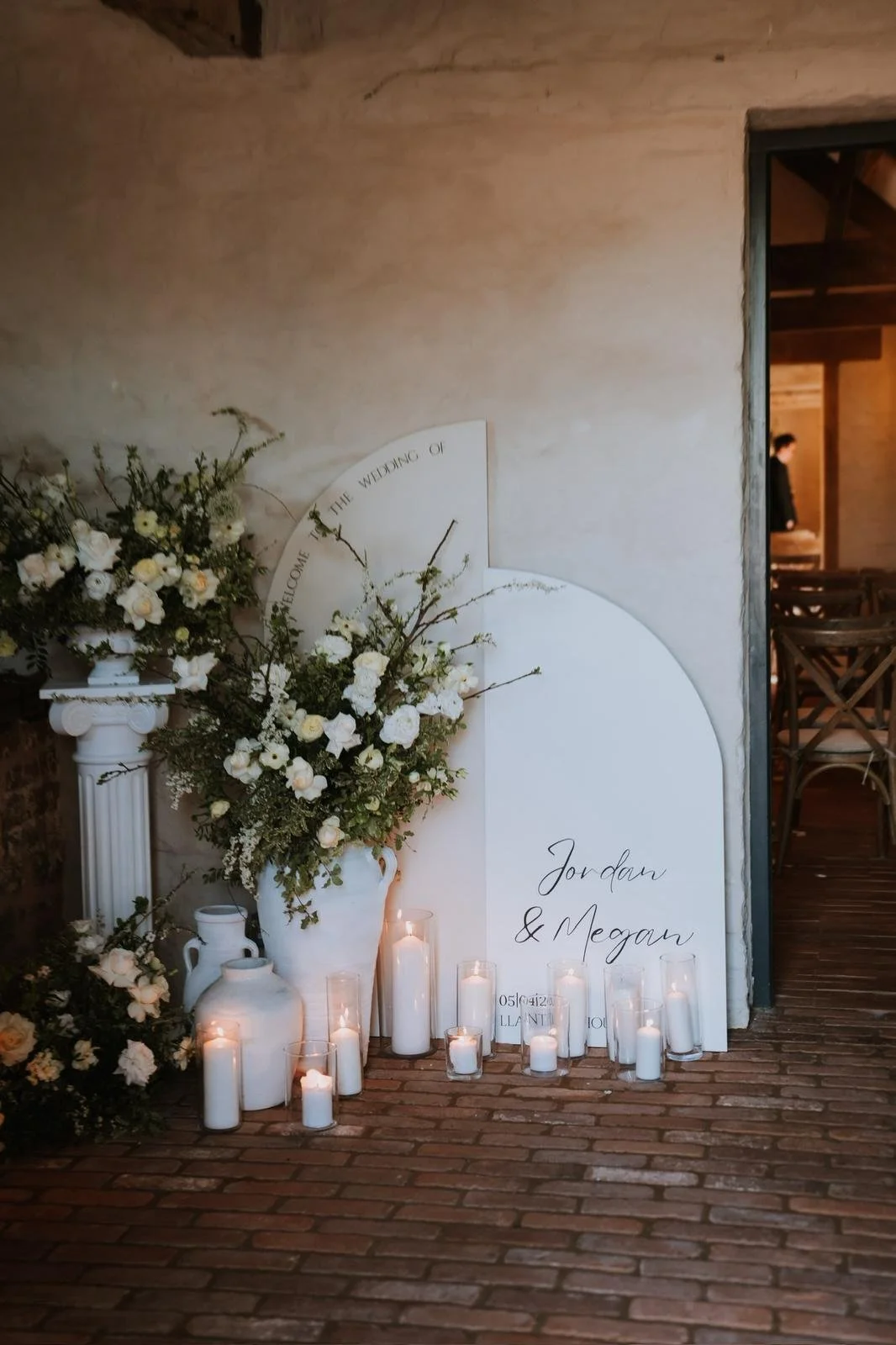Wedding decoration with white flowers in vase, candles, and signs with the names Jordan and Megan, on a brick floor with a doorway in the background.