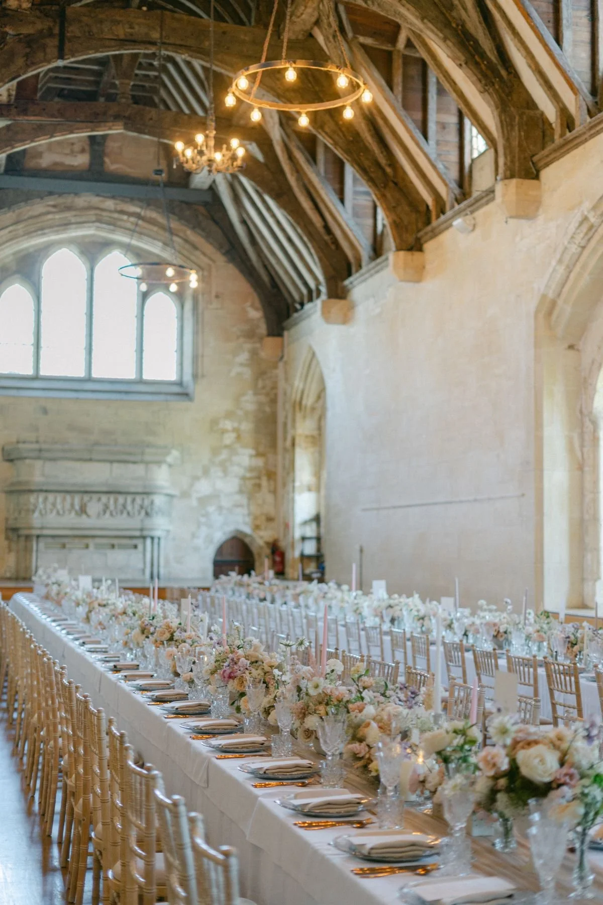 Long banquet table decorated with white and pastel floral centerpieces and candles inside a historic stone building with high arched windows and wooden ceiling beams.