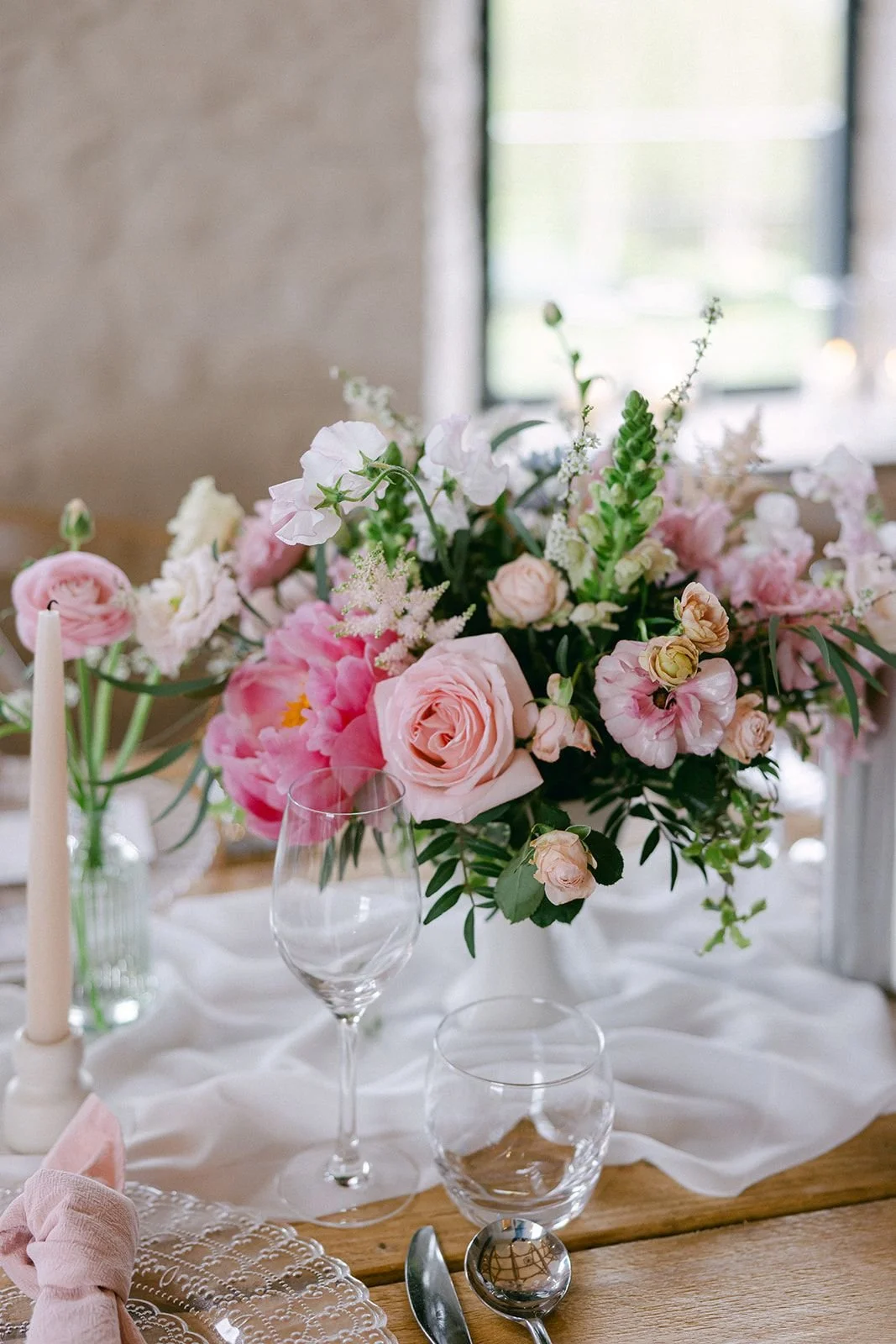 A floral centerpiece with pink and white flowers on a table with glassware and a white tablecloth, near a window.