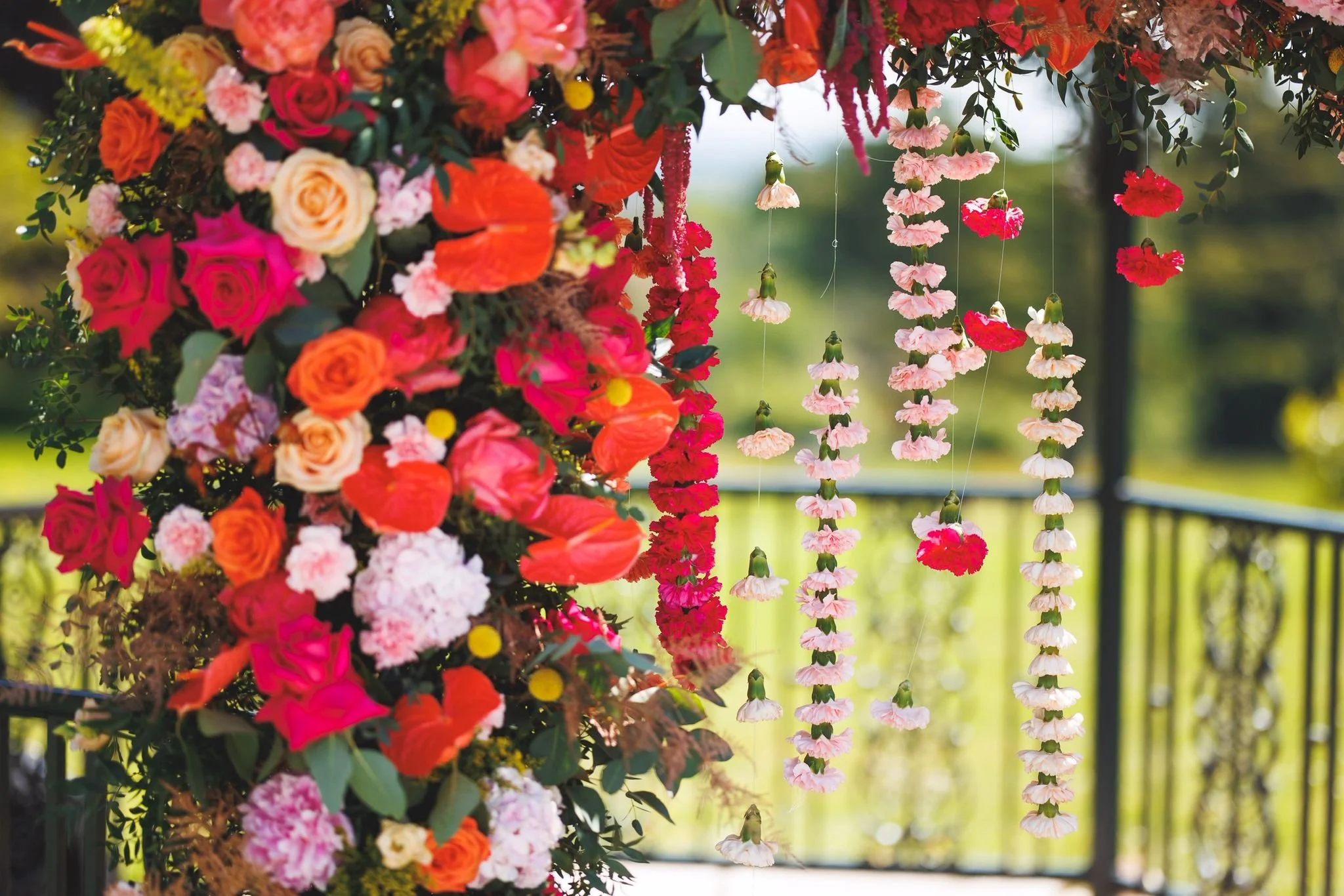 Colorful floral arrangement of pink, orange, red, and cream flowers with hanging pink and white garlands in the background on an outdoor balcony.