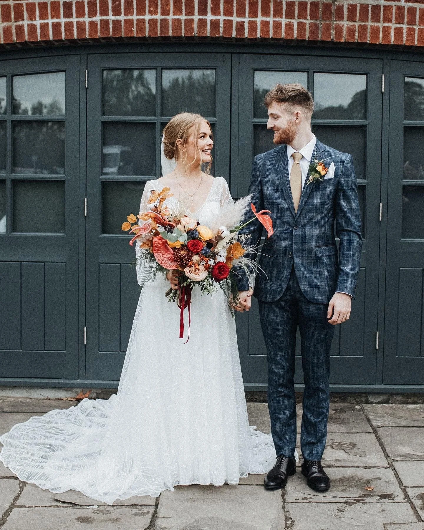 A bride and groom holding hands, smiling at each other, standing outside in front of a dark green door. The bride is wearing a white wedding dress and holding a bouquet of colorful flowers. The groom is wearing a blue checked suit with a white shirt,