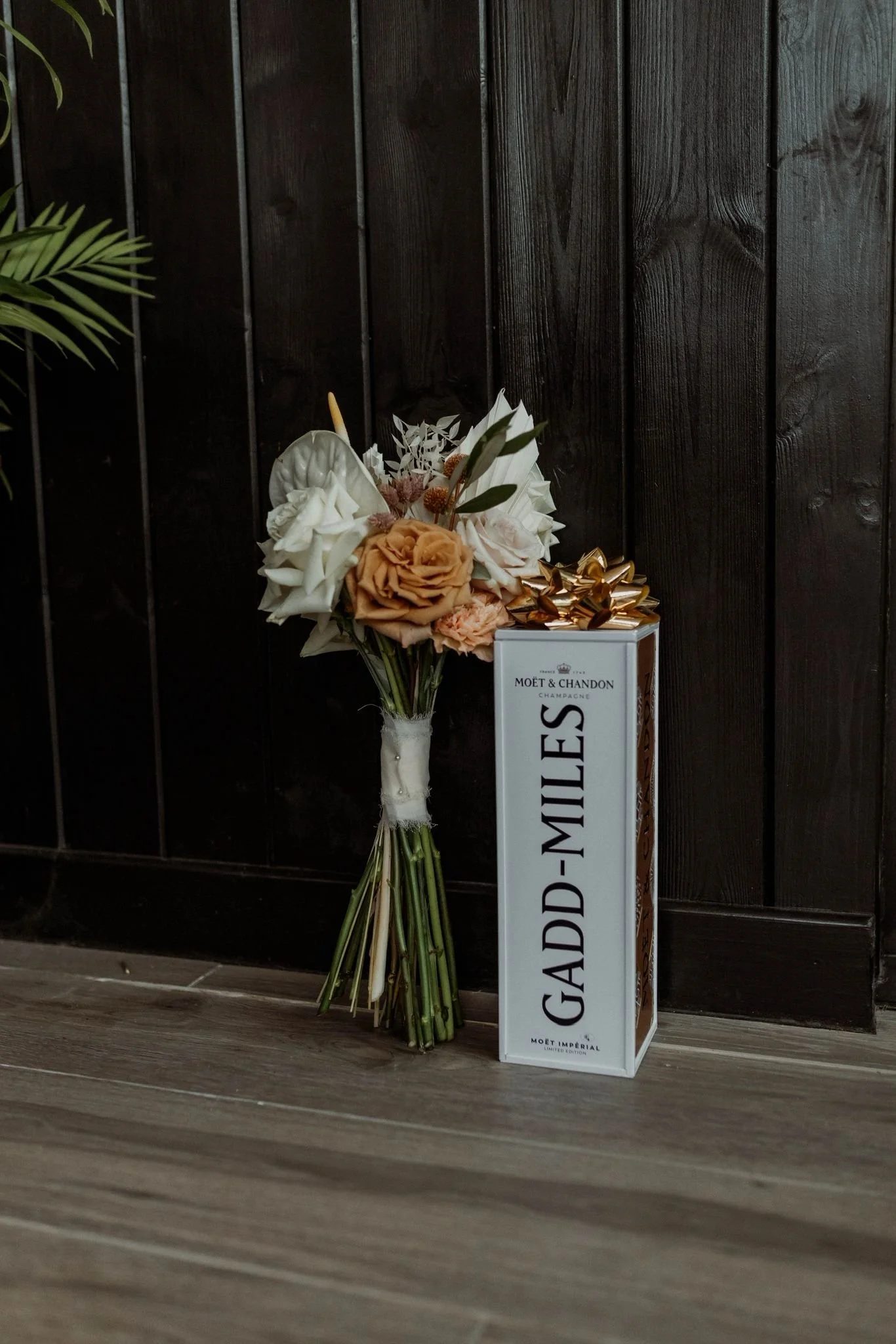A bouquet of flowers leaning against a dark brown wooden wall, next to a box of Moët & Chandon champagne labeled "GADD- miles" with a gold ribbon bow on top.