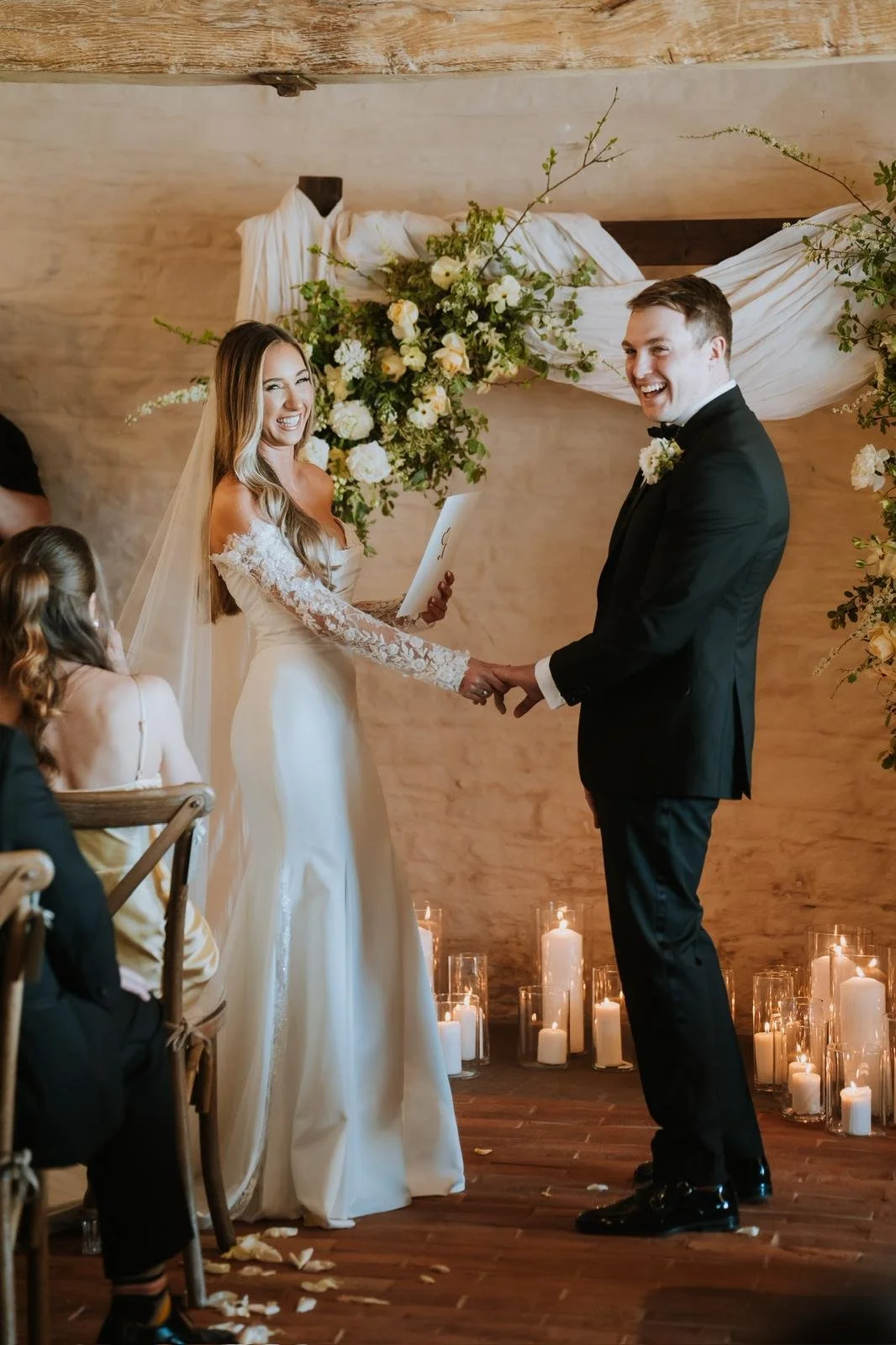 A bride and groom exchanging vows during a wedding ceremony, holding hands and smiling at each other, with candles and floral decorations in the background.