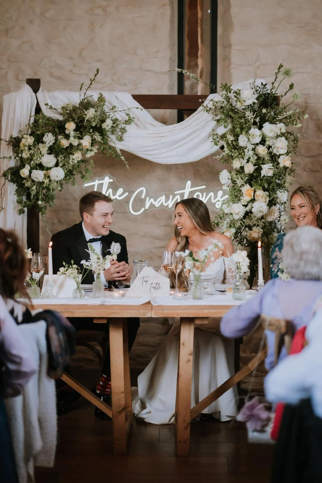 A wedding reception with a bride and groom sitting at a table, smiling and talking, with a bridesmaid beside them. The backdrop has floral arrangements and a neon sign that says 'The Crantone'.