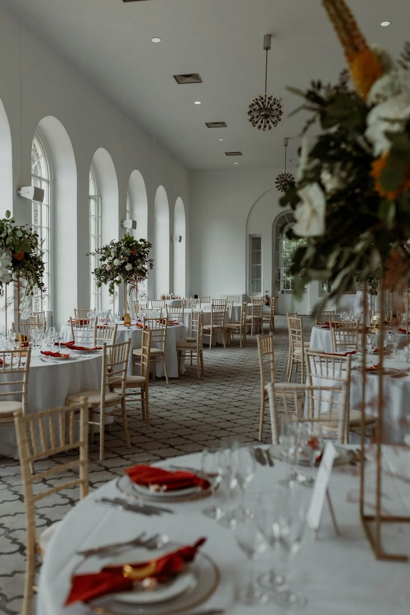 Elegant event space with round tables dressed in white tablecloths, arranged with glassware, silverware, and red napkins. Tall floral centerpieces with white and green flowers decorate the space, which features arched windows allowing natural light,