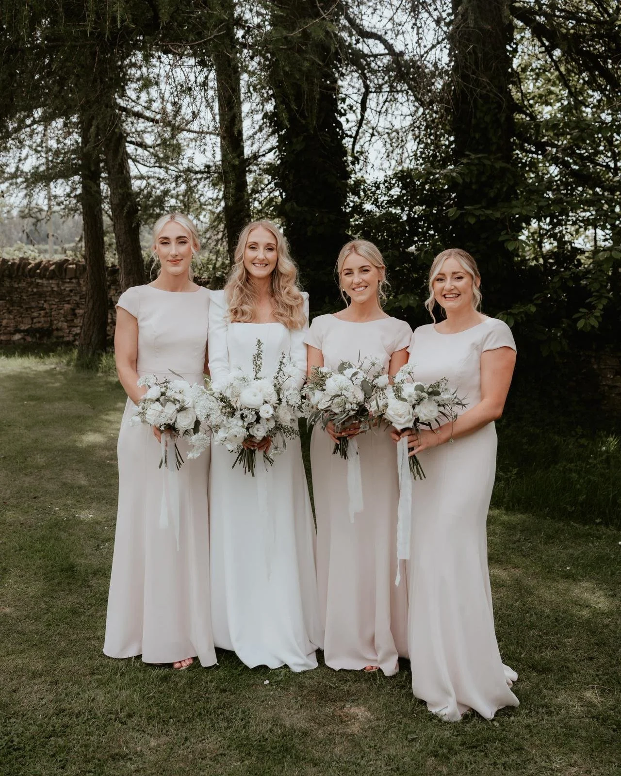Four women dressed in white, standing outdoors with greenery and trees in the background, holding white floral bouquets.
