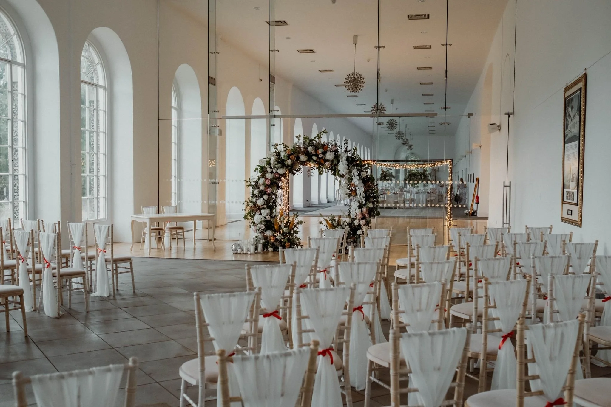 Wedding ceremony setup inside a bright hall with large arched windows, rows of chairs with white covers and red bows, a floral arch at the front, and a mirrored wall reflecting the decorated space.