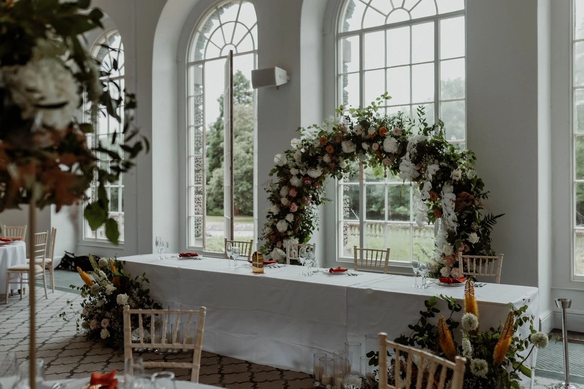 Elegant banquet table with floral arrangements, set in front of large arched windows in a bright room.