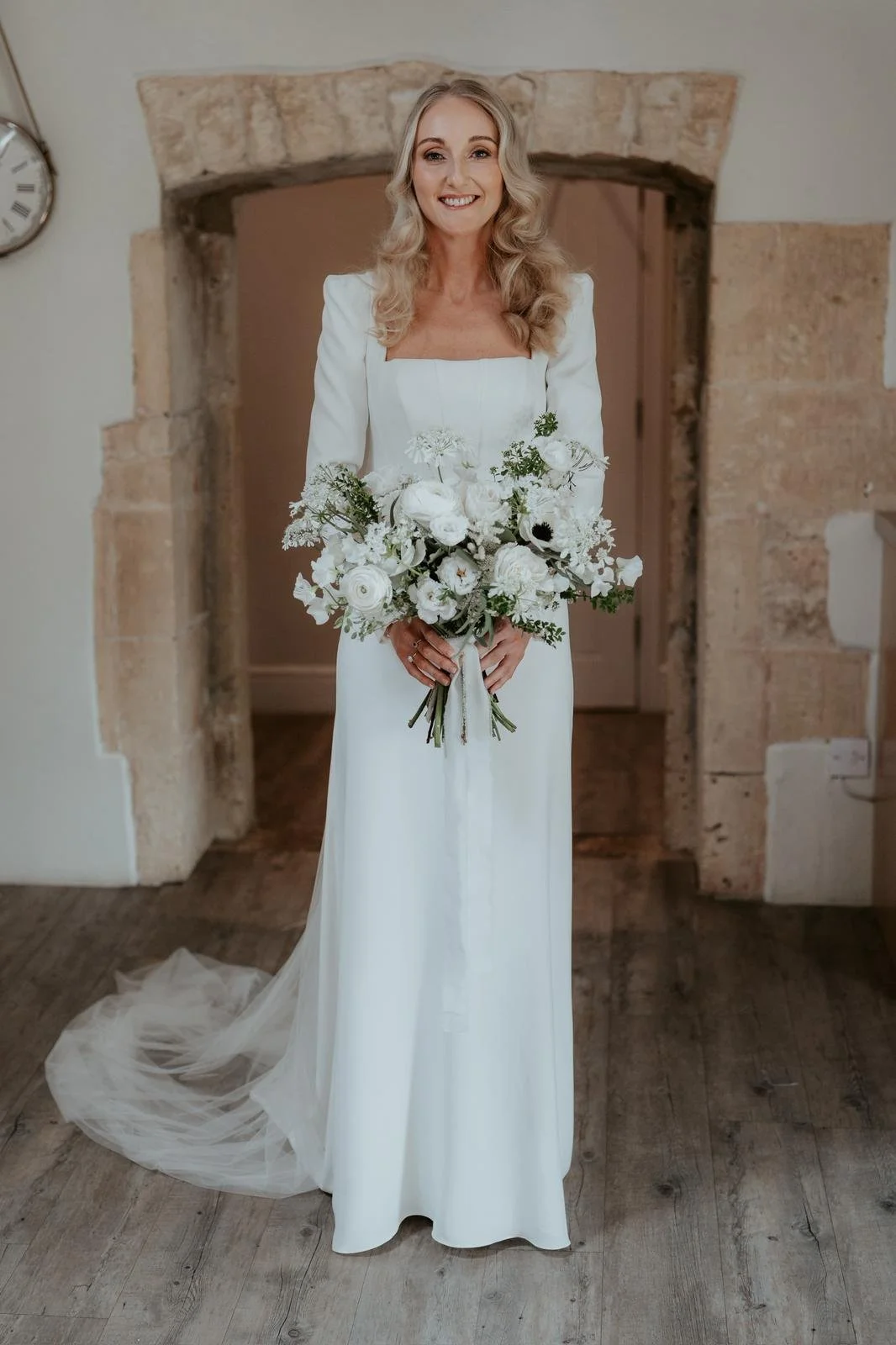 A smiling woman in a white wedding dress holding a bouquet of white flowers, standing indoors in front of a stone wall and wooden floor.