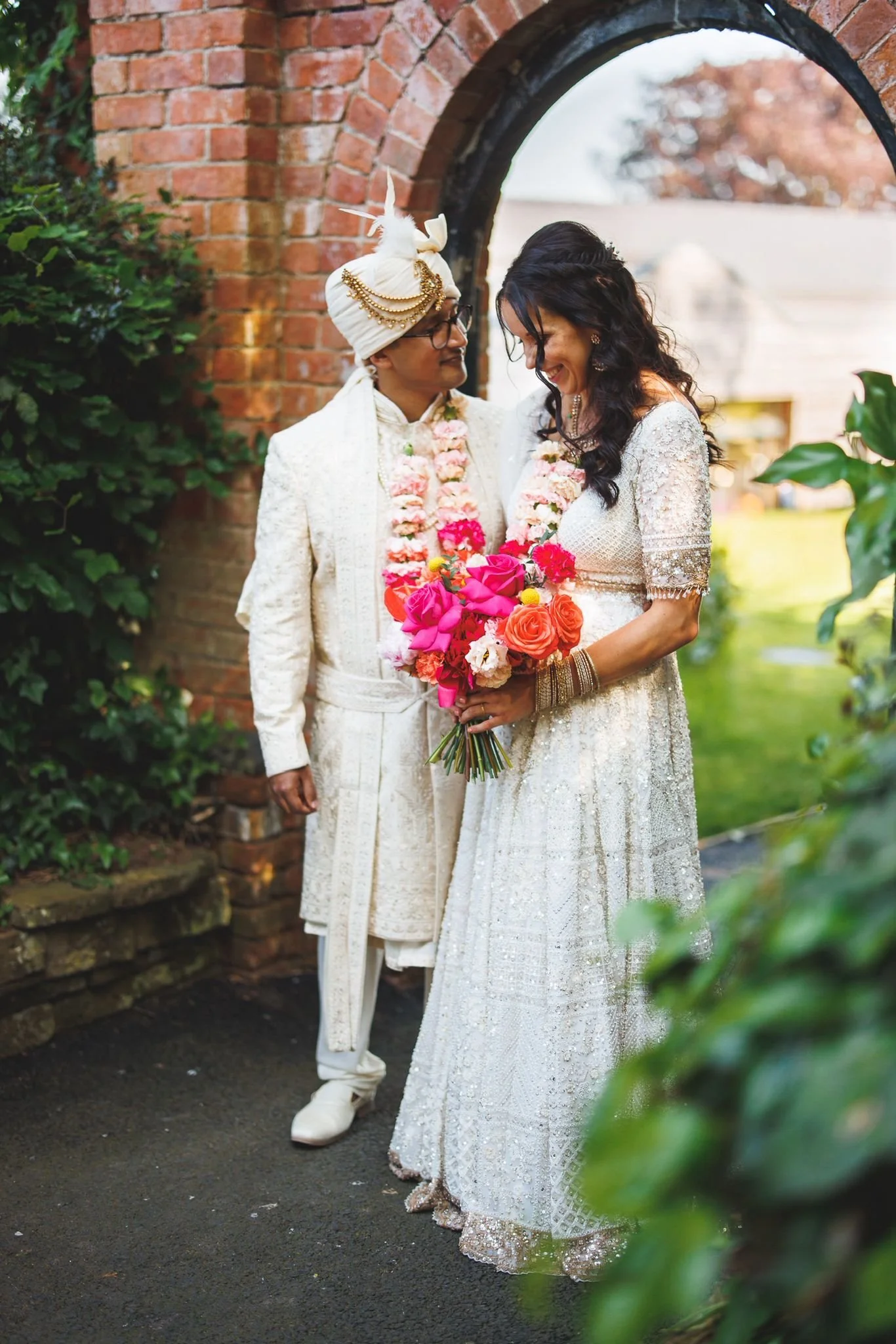 A bride and a groom in traditional Indian wedding attire standing under an arched brick doorway. The bride holds a vibrant bouquet of pink, orange, and white flowers and looks down smiling. The groom, dressed in white with intricate embroidery and a