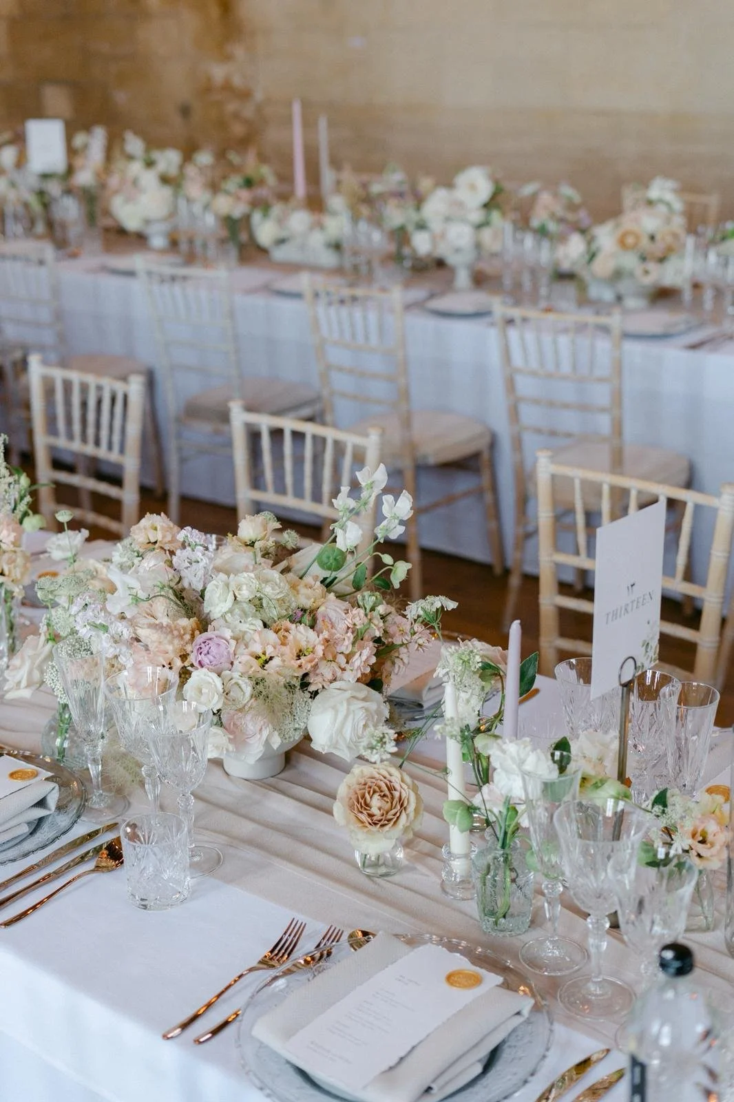 Elegant wedding table decorated with a pastel floral centerpiece, candles, glassware, plates, silverware, and a wedding menu, with empty chairs around.