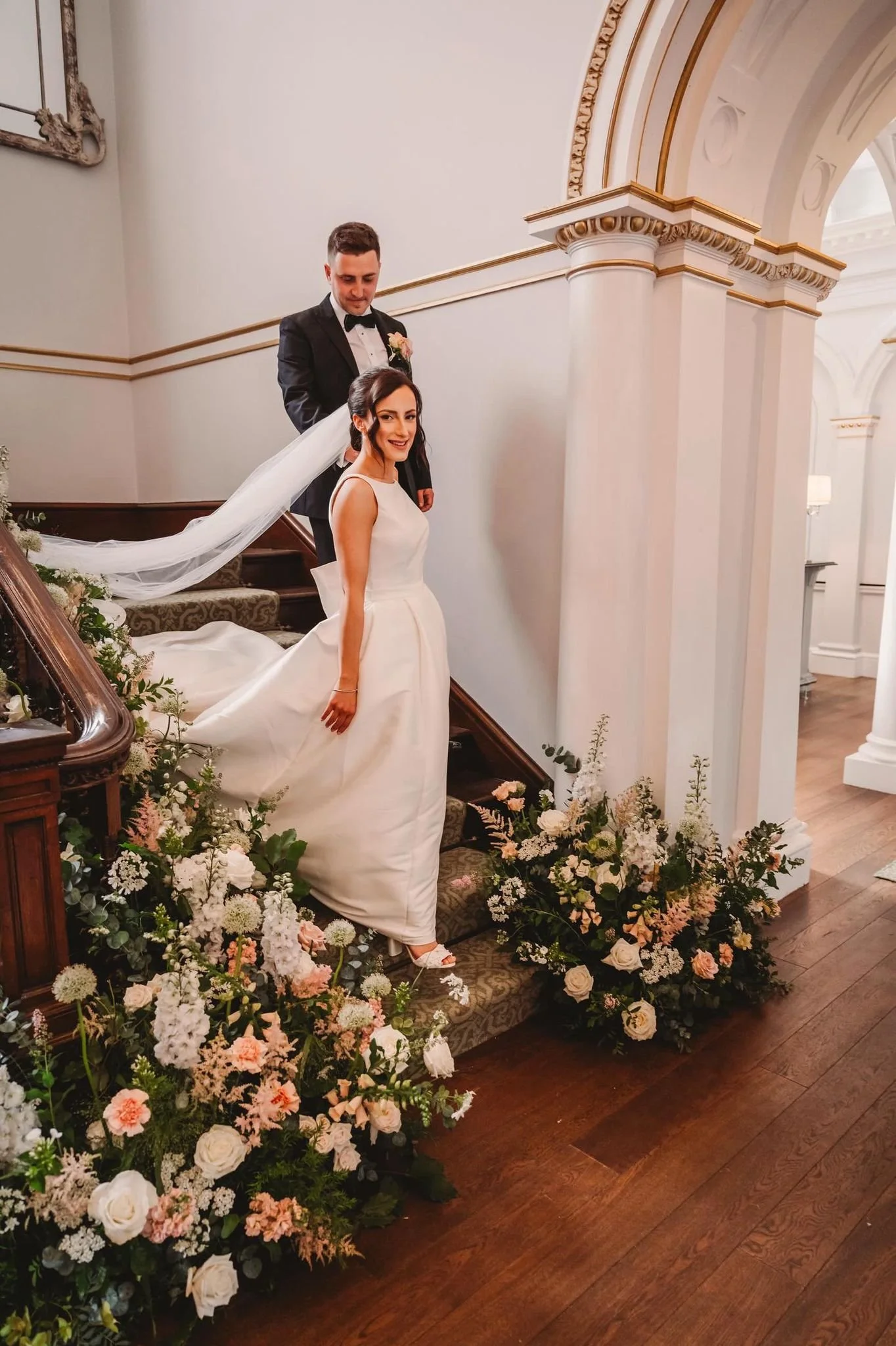 A bride in a white wedding gown and veil stands at the bottom of a staircase decorated with white and soft pink flowers. A groom in a black tuxedo stands behind her on the stairs. The setting is an elegant, well-lit interior with white walls and clas
