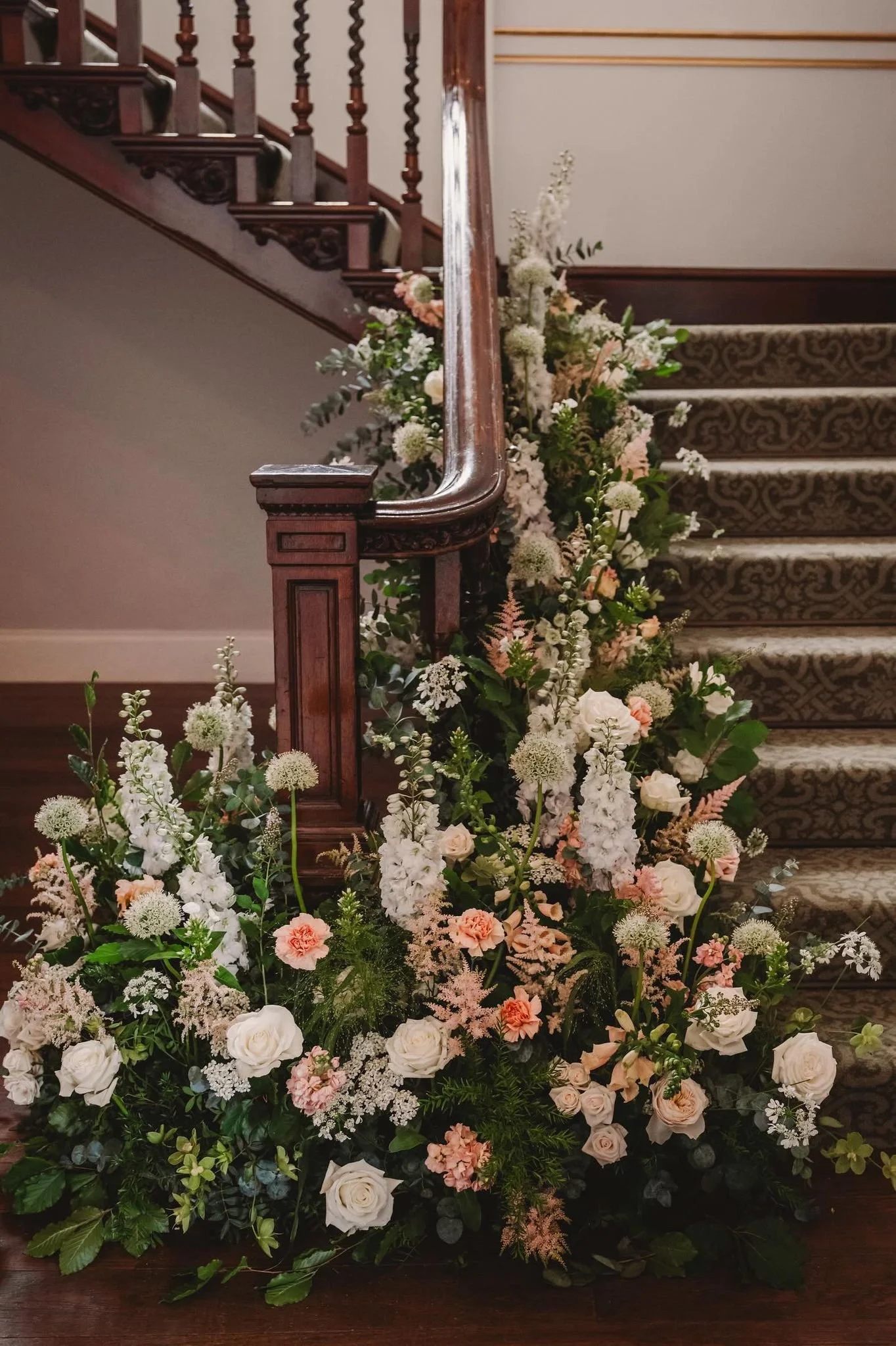 A floral arrangement with white, pink, and peach flowers at the base of a wooden staircase with a dark, patterned carpet.