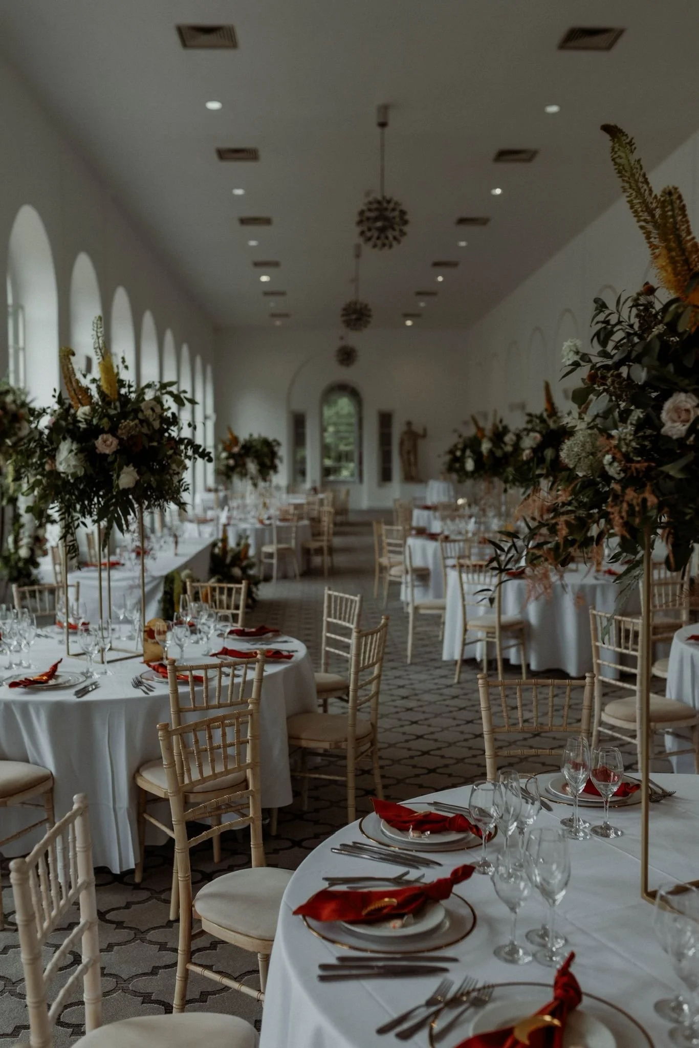 Elegant banquet hall decorated with white tablecloths, tall flower centerpieces, and gold-chained chairs, set for a formal event.