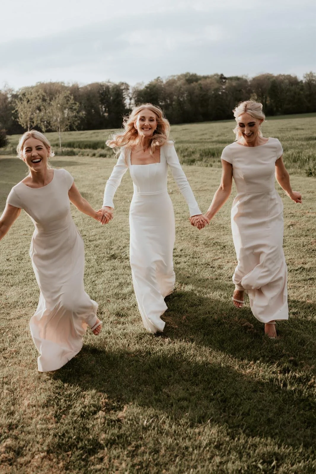 Three women in white dresses holding hands and running through a grassy field, smiling and enjoying the moment.