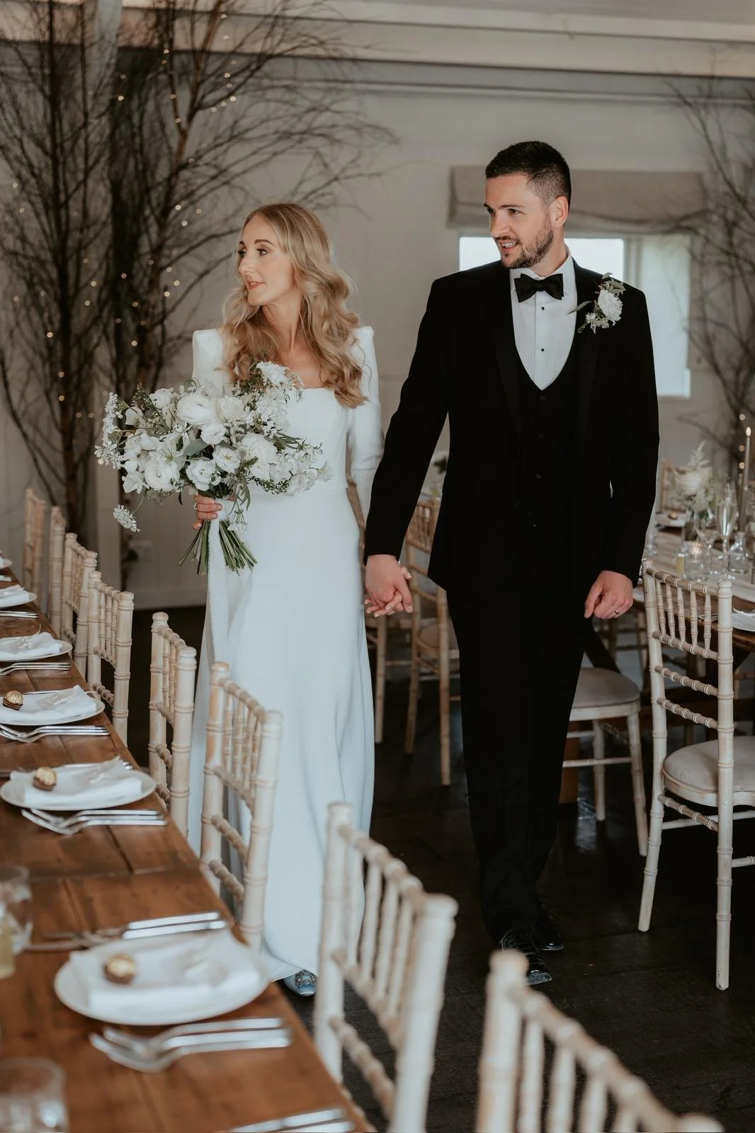 A bride and groom walking hand in hand at their wedding reception, with the bride holding a bouquet of white flowers, in a decorated event space.