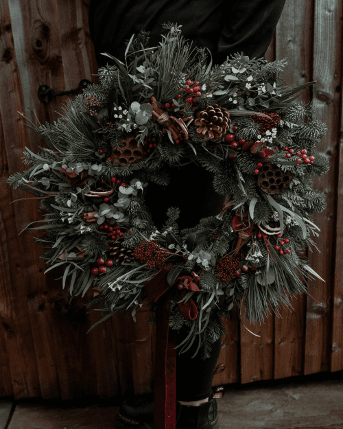 Person holding a holiday wreath made of pine branches, pinecones, red berries, and greenery against a wooden background.