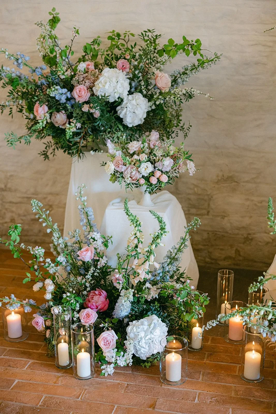 Elegant floral arrangement with pink, white, and purple flowers, surrounded by lit candles in glass holders, on a brick floor against a beige wall.
