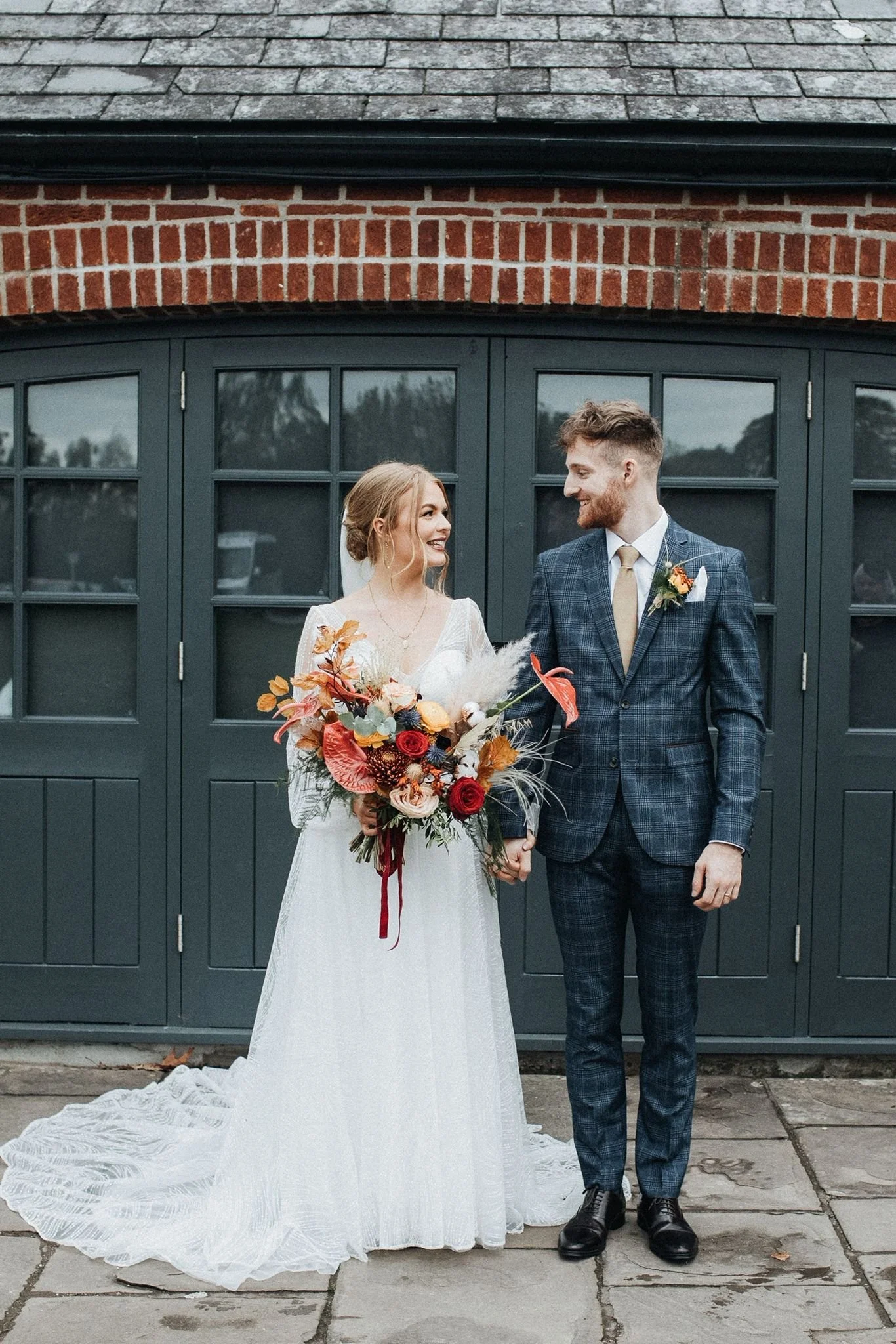 A bride and groom stand outside, holding hands and looking at each other. The bride wears a white wedding dress and holds a colorful bouquet, while the groom wears a blue plaid suit with a tan tie. They are in front of a building with dark green wind