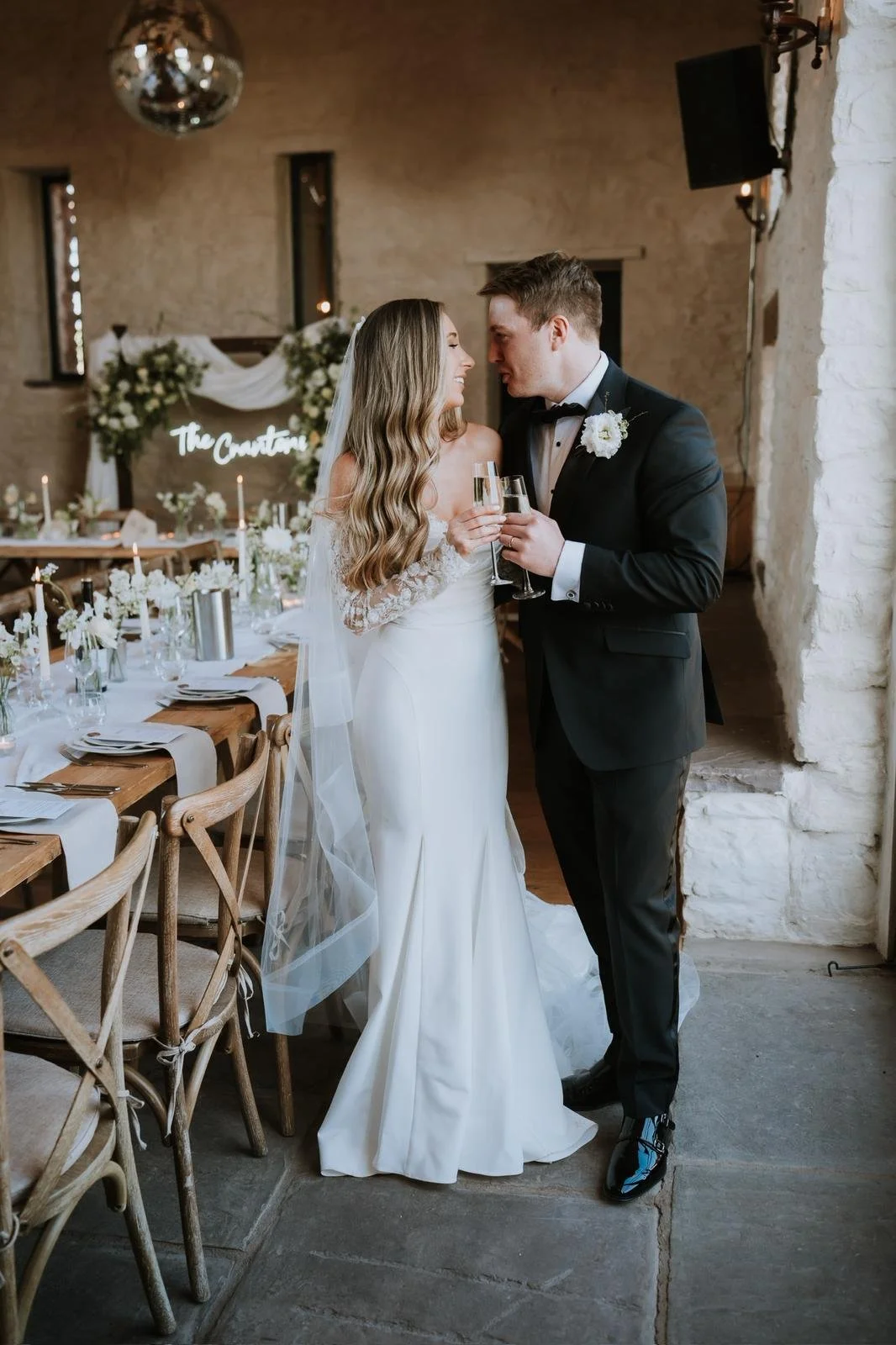 A newlywed couple raising glasses of champagne during their wedding reception in a decorated rustic venue with a long dining table and floral arrangements.