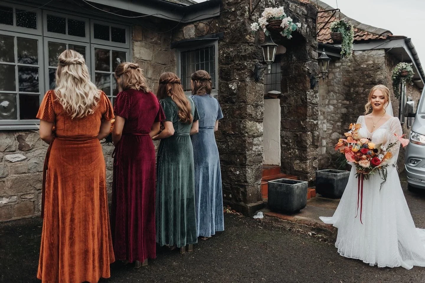 A bride holding a bouquet of flowers stands outside in front of four bridesmaids, all facing away from the camera, in front of a stone building with hanging flower baskets.