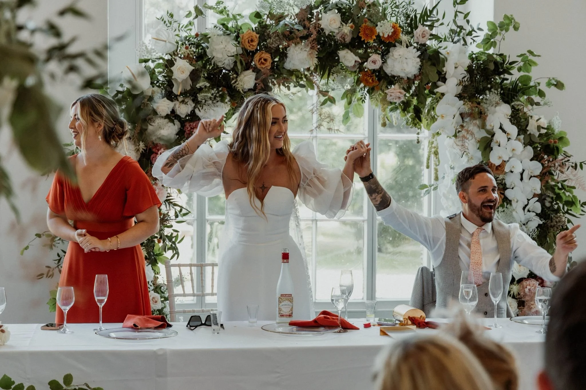 A bride in a white wedding dress and a woman in an orange dress stand on either side of a groom in a vest, seated at a decorated table with floral arrangements and glasses, inside a bright room with large windows and floral arch background. They appe