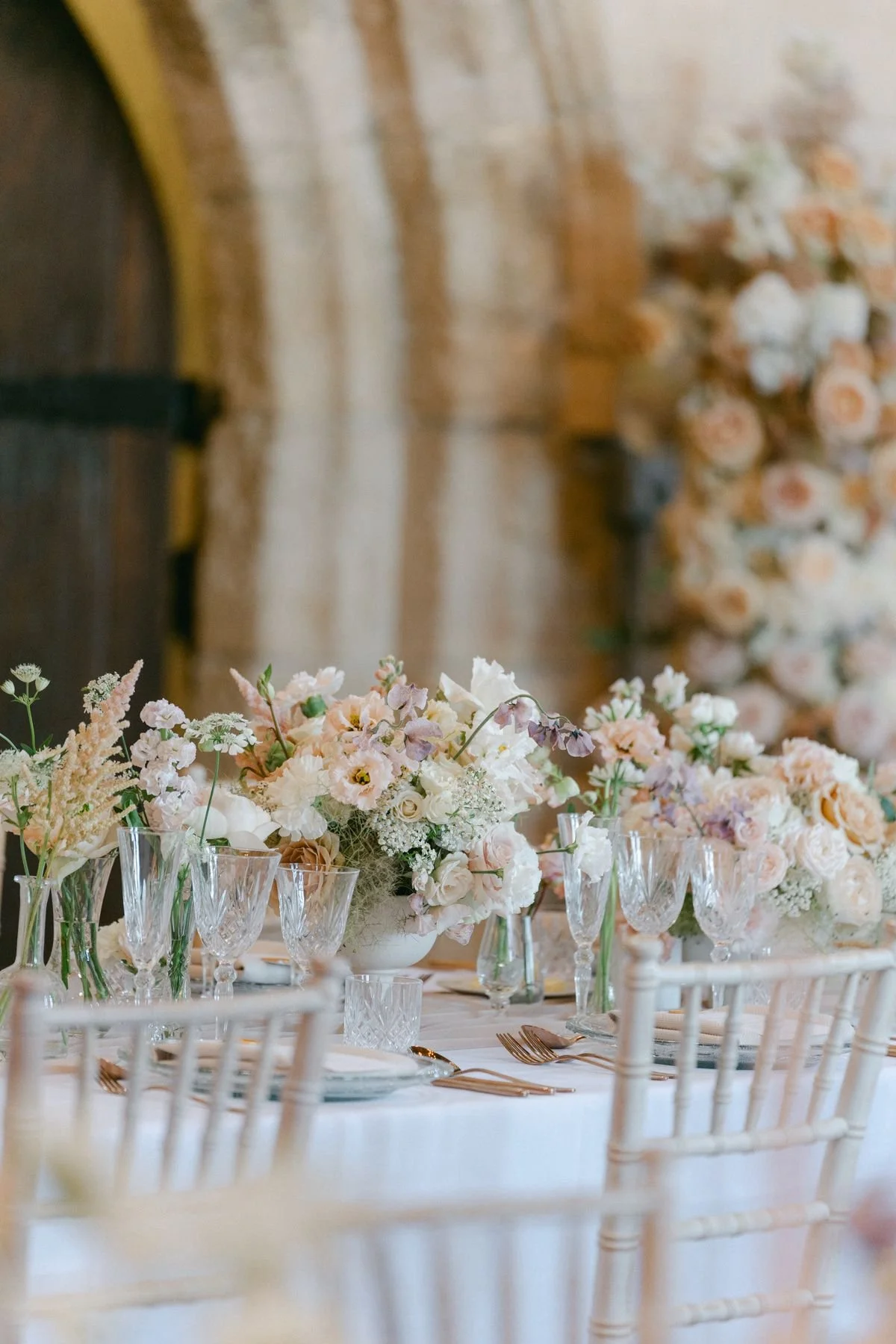 Elegant table setting with white and blush floral centerpieces, crystal glassware, and gold flatware in a rustic venue with exposed brick and wooden accents.